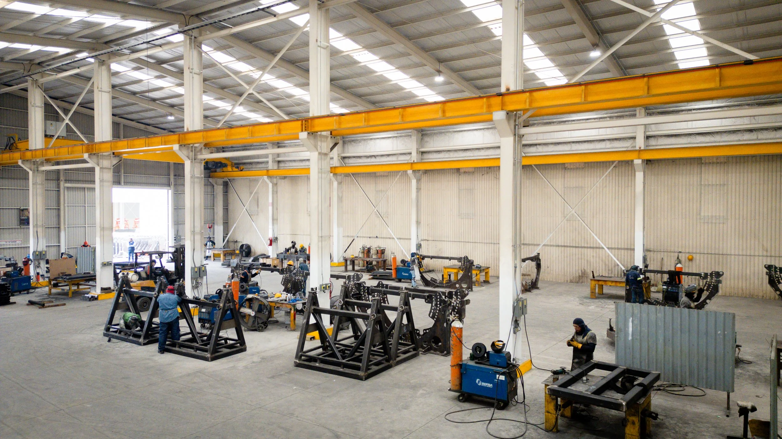 Interior de una fábrica con maquinaria y trabajadores en procesos de fabricación, estructura metálica y techos altos.