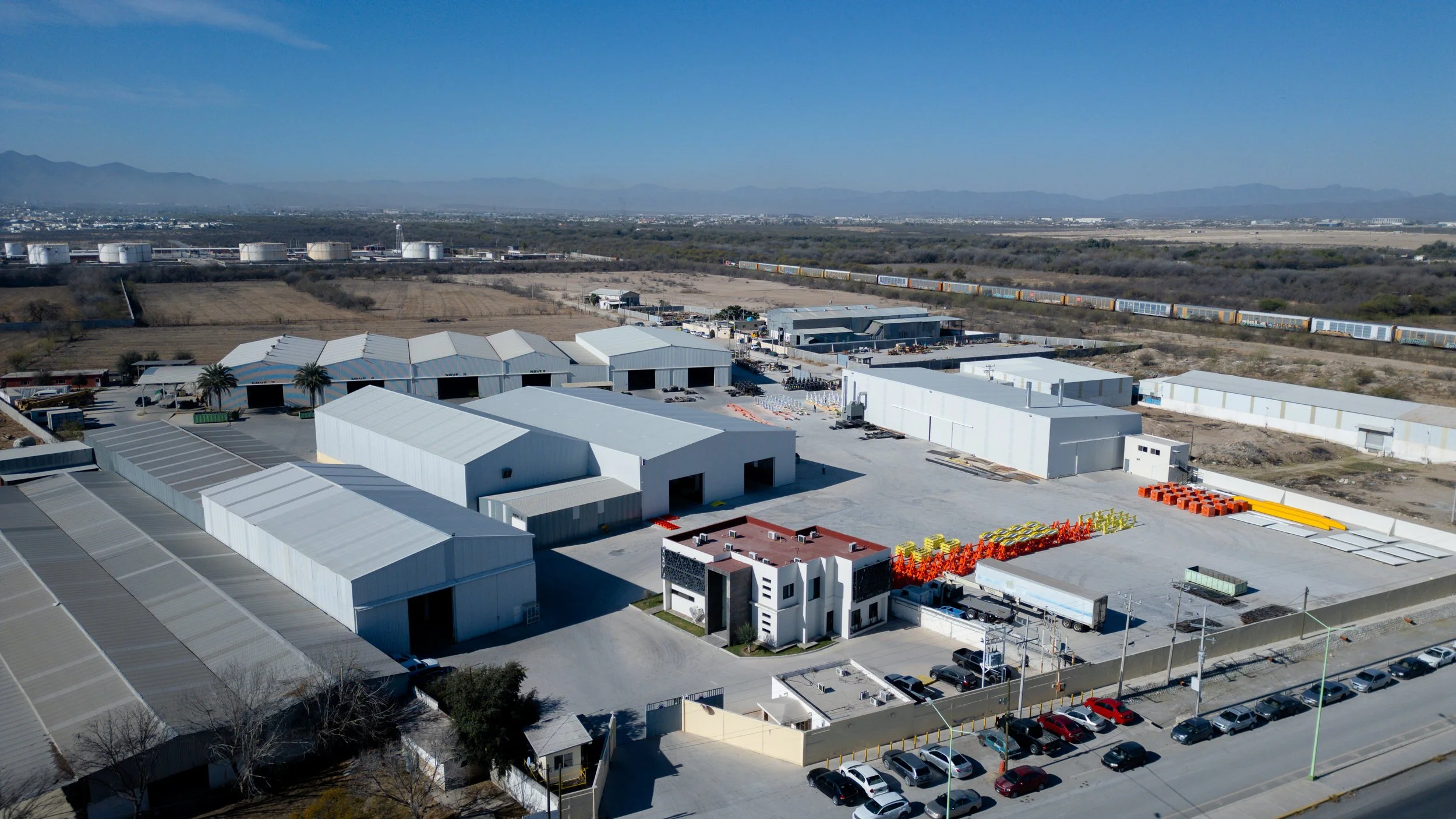 Vista aérea de una zona industrial con edificios blancos, almacenamiento y tren de carga en el fondo, en un paisaje árido con montañas al horizonte.