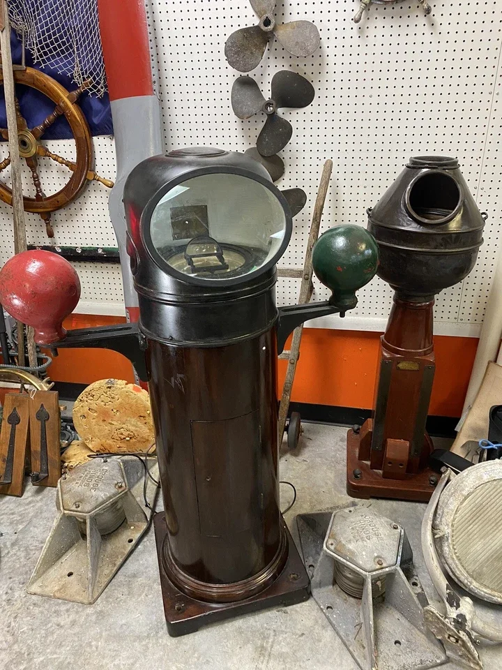Vintage cockpit simulator with a black cylindrical stand, chrome spheres on either side, and a large glass-covered control panel with a steering wheel inside, set in an antique display with other maritime and mechanical items in the background.