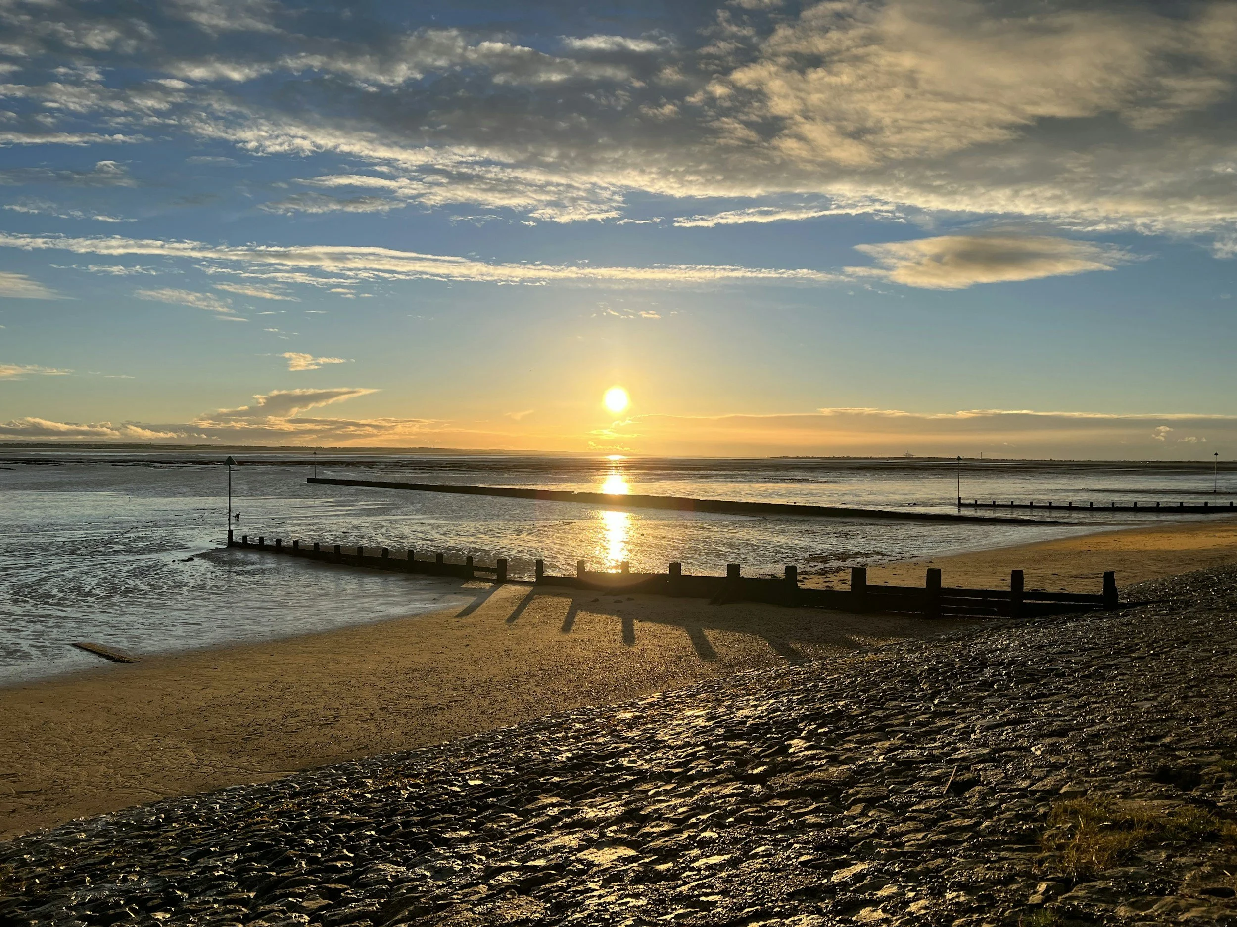 Sunset over a beach with calm water, a wooden pier, and scattered clouds in the sky.