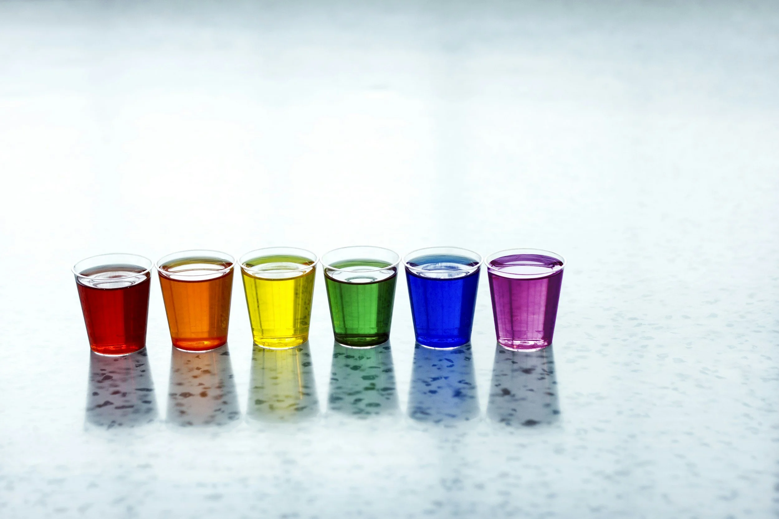 Six shot glasses lined up in a row with liquids of different colors in them, forming a rainbow.  Left to right: Red, orange, yellow, green, blue, purple