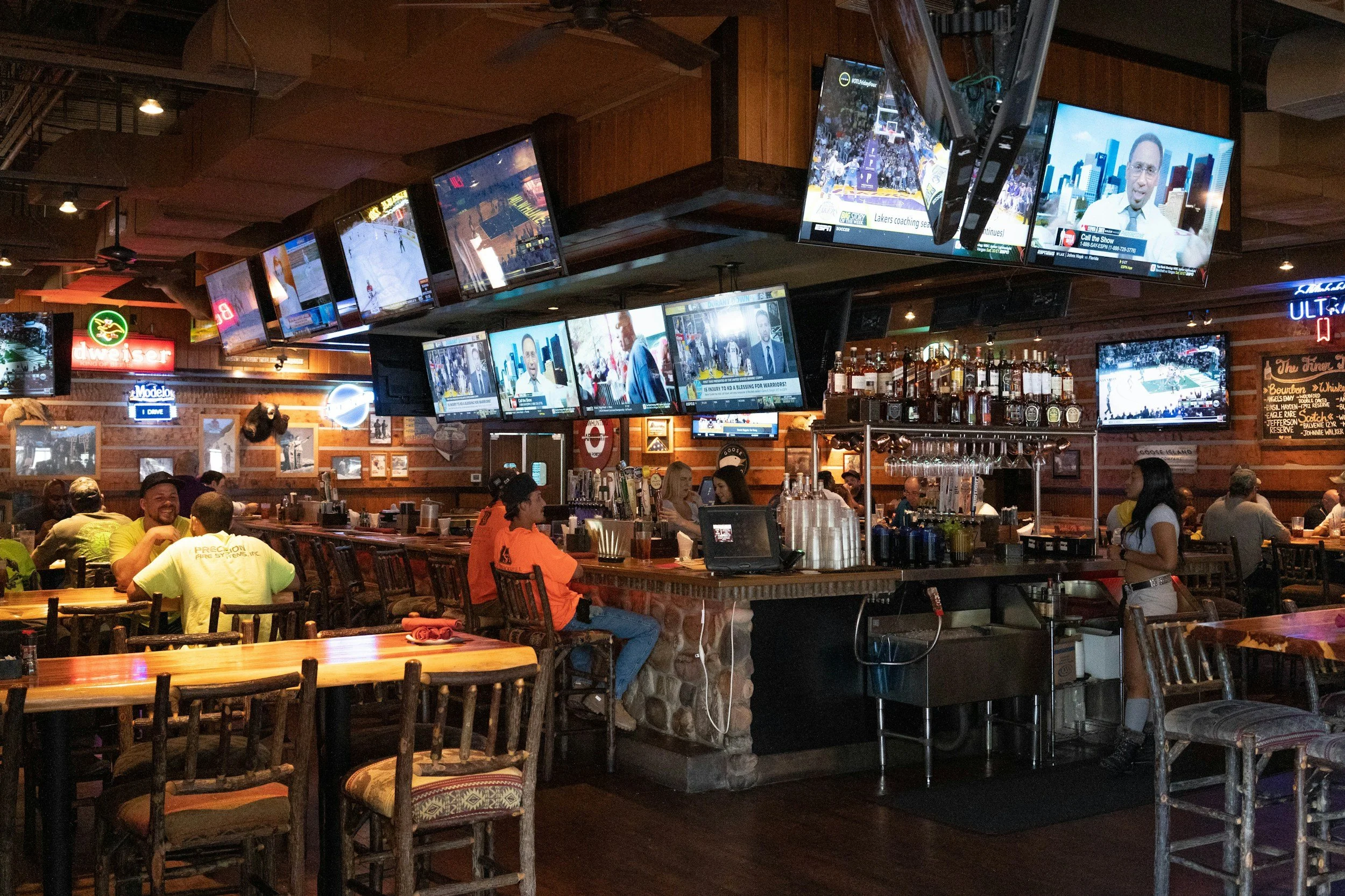 The inside of a corporate bar with people at tables and the bar.  It is a rustic interior with neon signs and many TVs