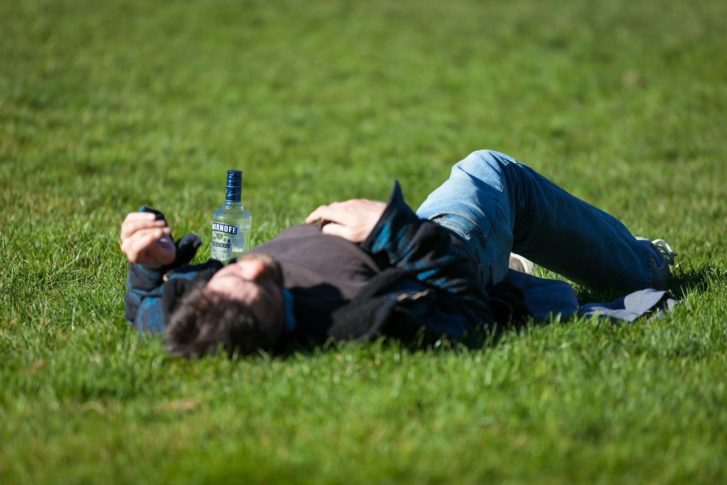 young man passed out in grassy field with a bottle of liquor next to him.  Don't pass out in the grass, that's where the ants are.
