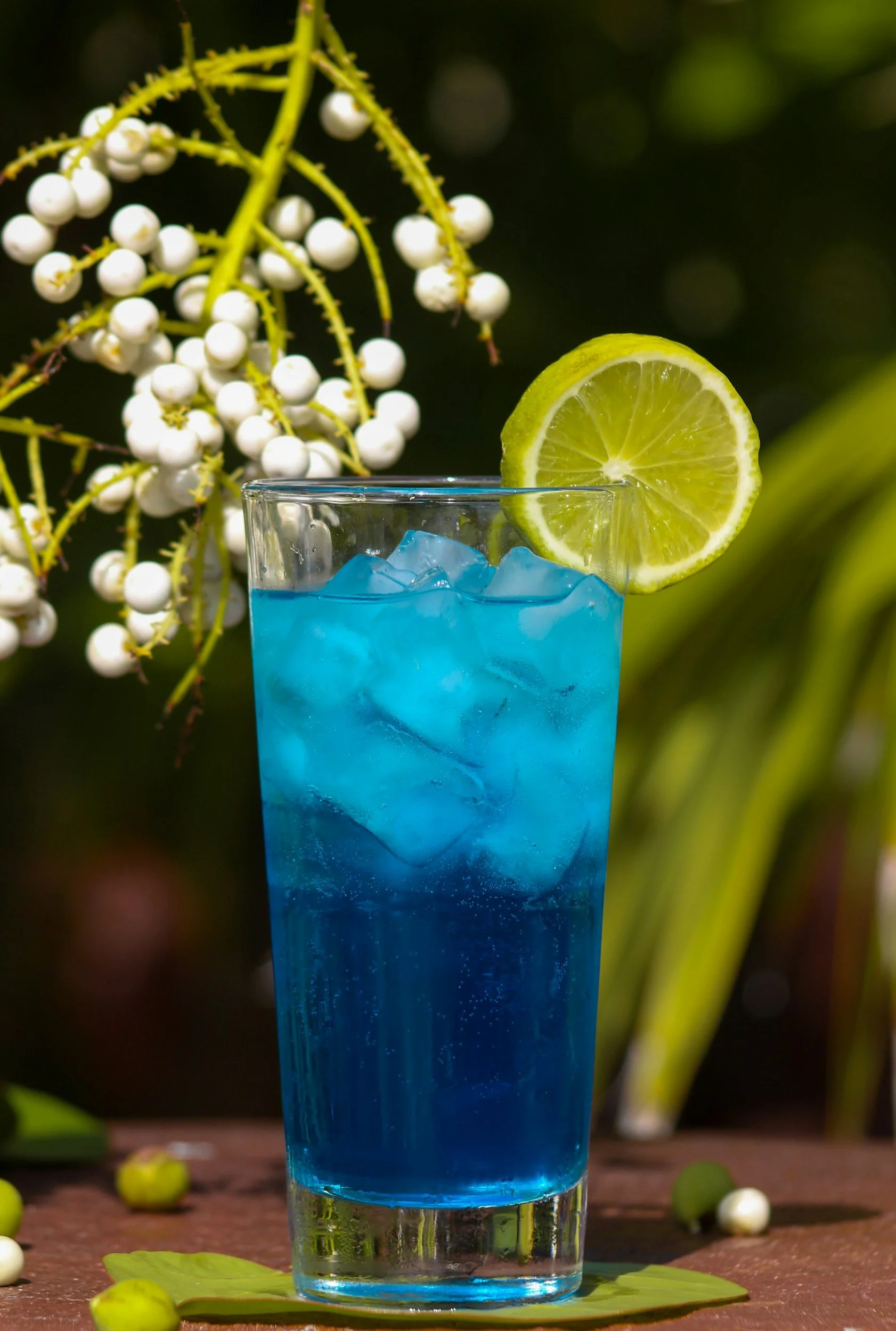 Side view of a blue cocktail in a pint glass with a lemon wheel on the rim.  There are white flowers and greenery in the background