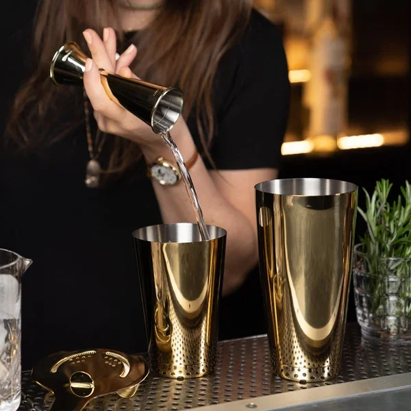 A woman using a jigger to pour liquid into a set of gold plated shaking tins.