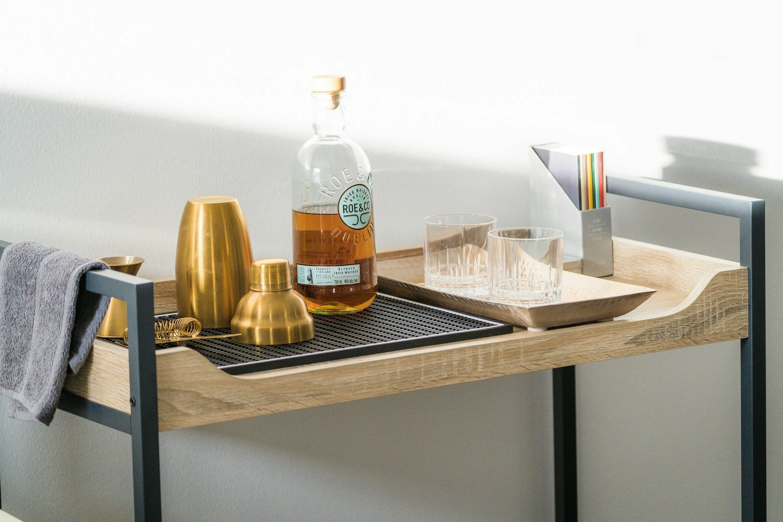 A wooden bar cart with a bottle of alcohol, shaker, and glasses