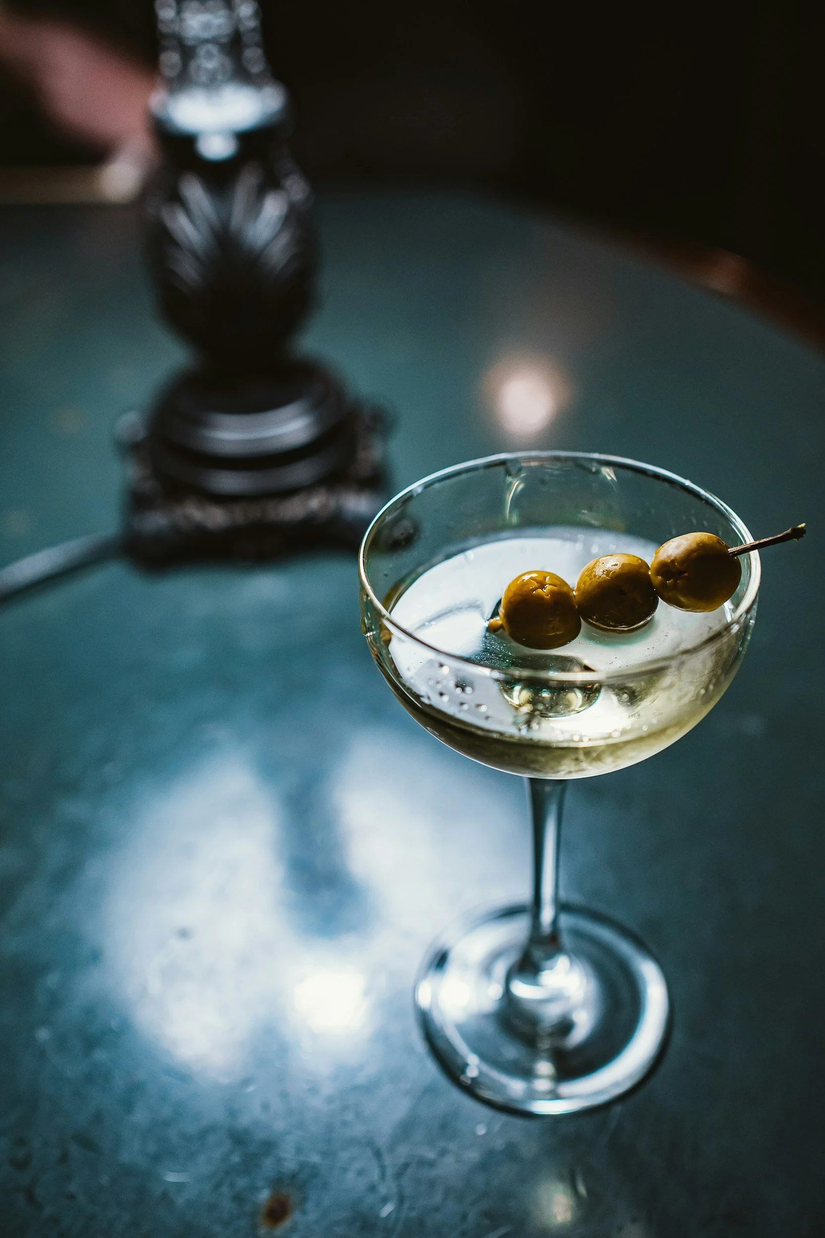 Martini in a coupe glass with three olives on a cocktail stick.  Background is dark blue and turquoise.