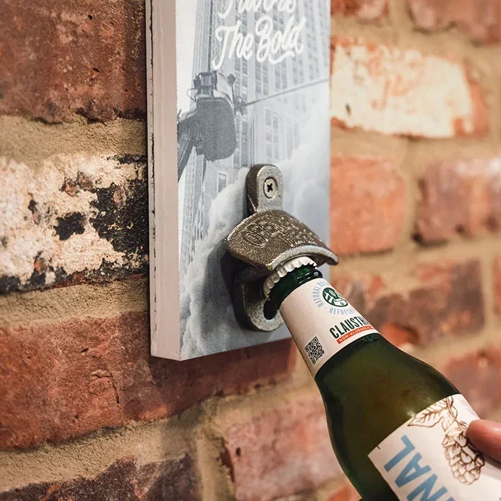 A metal bottle opener mounted to a wooden board.  It is set on a brick wall and being used to open a bottle of beer