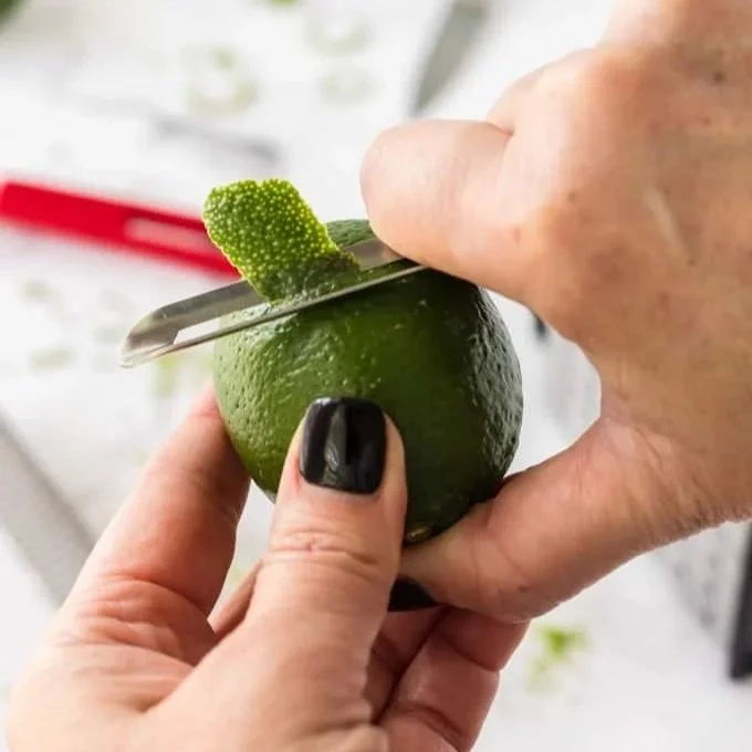 A woman with black nails holding a lime with one hand and using a side-hold peeler to lift off a half an inch of lime peel from the fruit.