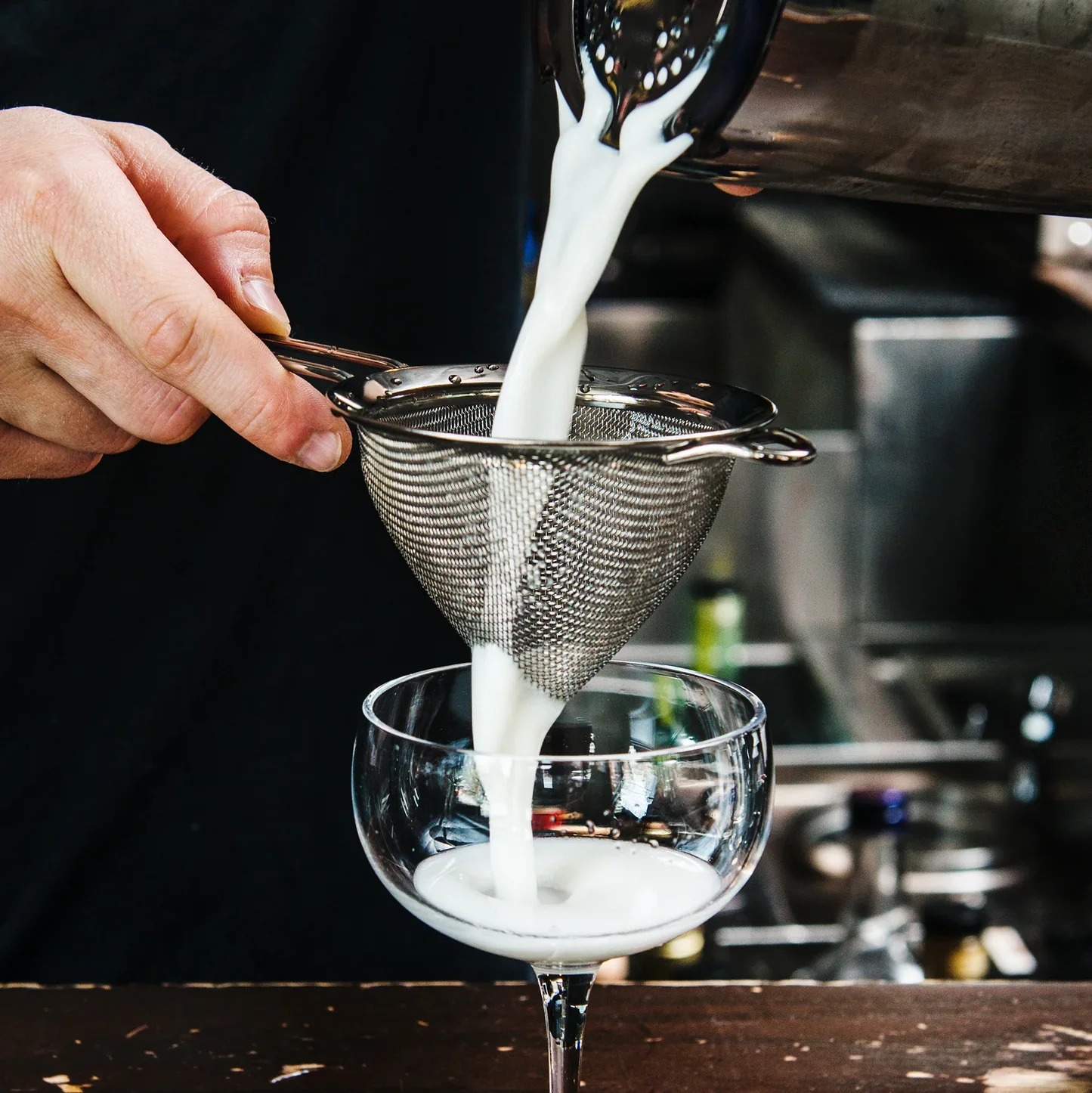 a man using a mesh strainer, a metal loop with a handle, with metal mesh hanging off of it in a cone shape.  The fine mesh catches finer pieces of ice and herbs