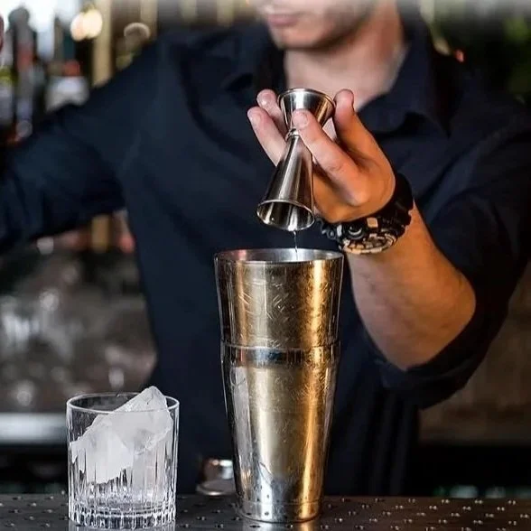 Bartender using a jigger to pour into a metal tin.  The jigger is two cones that are joined at the base, each holding a different volume of liquid.