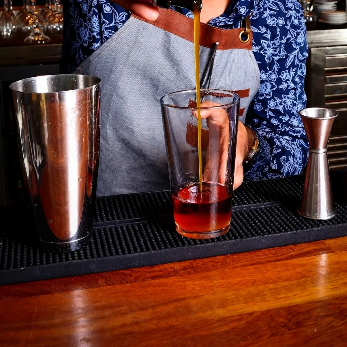 A bar using two long, thin bar mats.  there is a jigger, metal tin, and glass with liquid on the mat