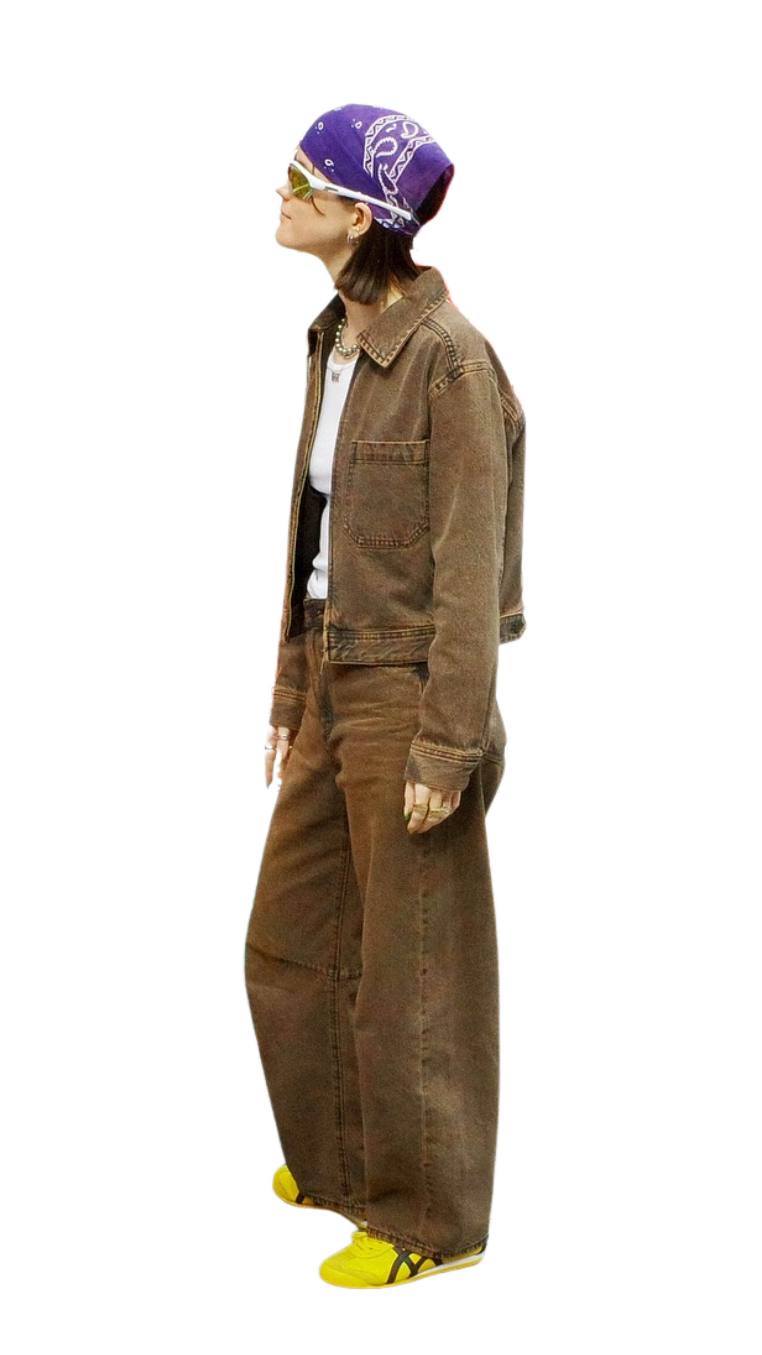 A person with shoulder-length dark hair, wearing a purple bandana, white sunglasses, a brown oversized denim jacket, brown wide-leg jeans, and yellow sneakers, standing in profile.