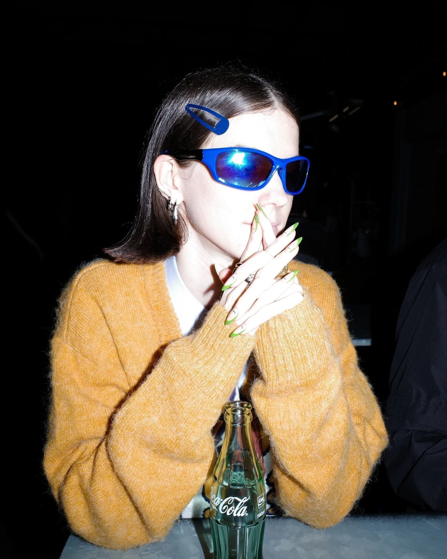 A woman with dark hair, wearing blue sunglasses and a yellow sweater, is sitting at a table with a Coca-Cola glass bottle in front of her. She is holding her hands near her face with fingers crossed, and there is a short glance at her profile. The background is dark and out of focus.