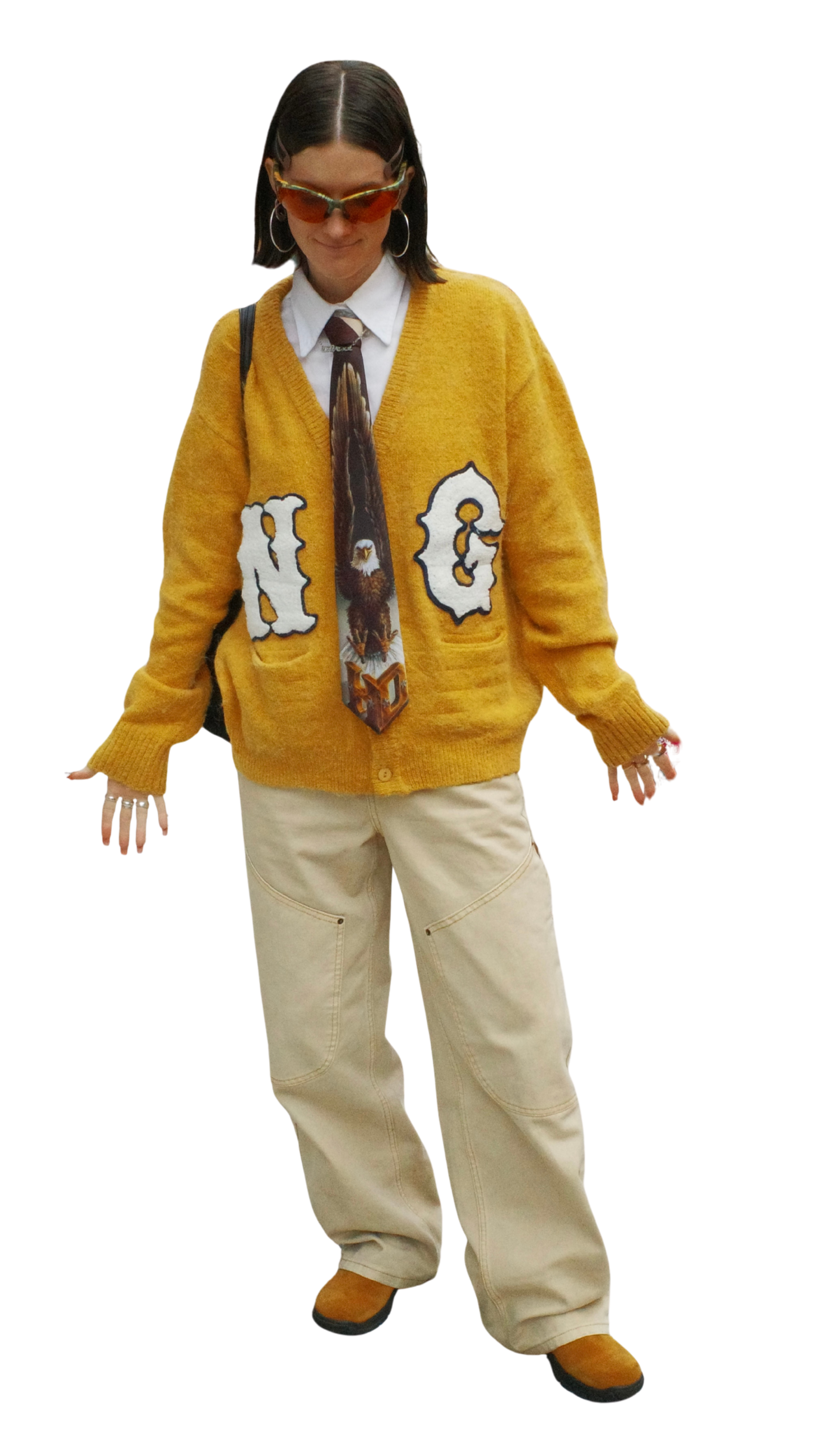 A woman wearing large orange sunglasses, hoop earrings, a mustard yellow cardigan with patches spelling 'NG', a white shirt, a patterned tie with an eagle image, beige cargo pants, and brown shoes, standing with a relaxed posture.