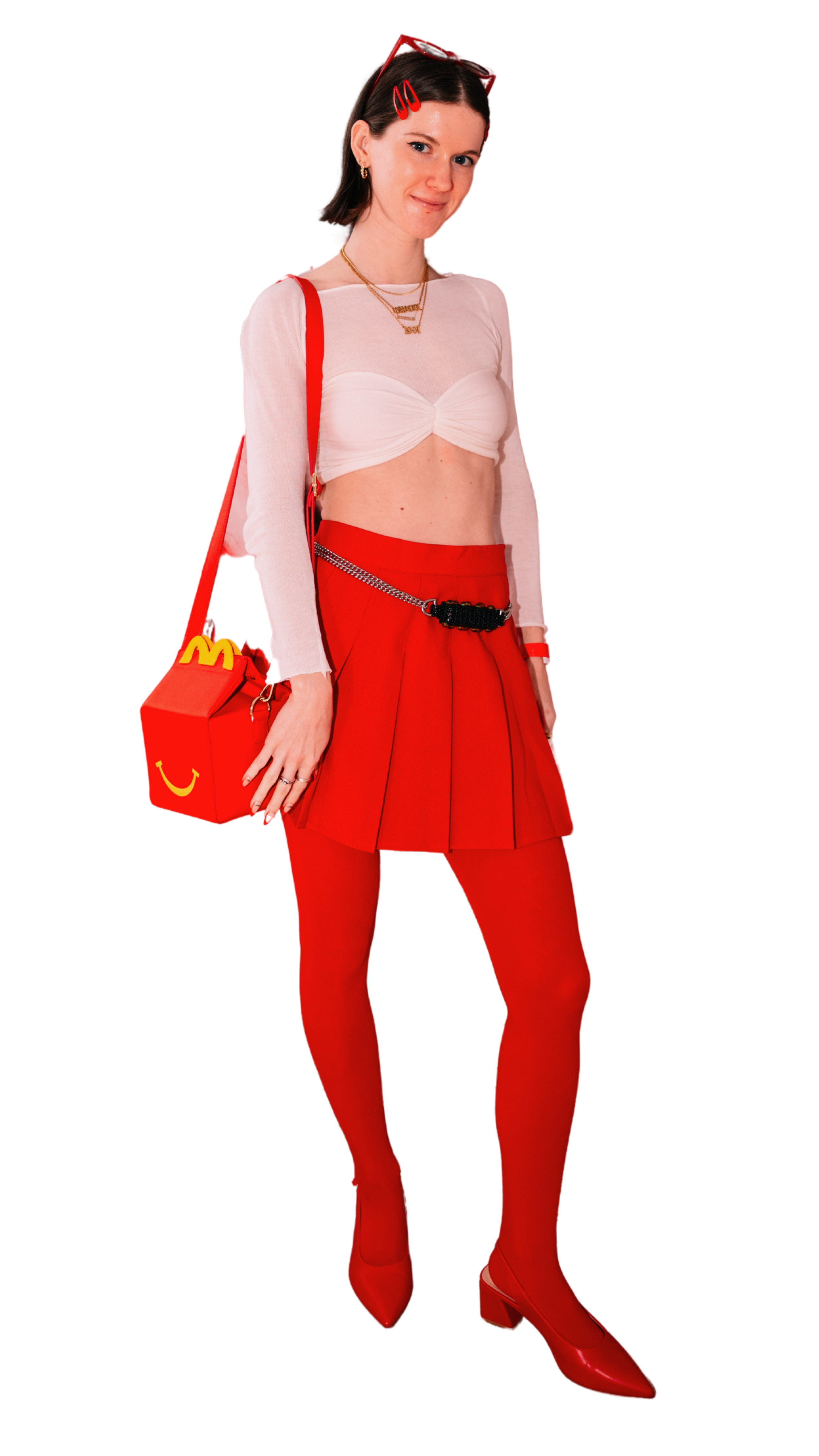 A woman with brown hair wearing red sunglasses on her head, gold earrings, layered gold necklaces, a white sheer long-sleeve top with a sweetheart bust, bright red high-waist pleated pants with a chain belt, red pointed-toe high-heeled shoes, and carrying a red handbag with and a yellow McDonald's logo.