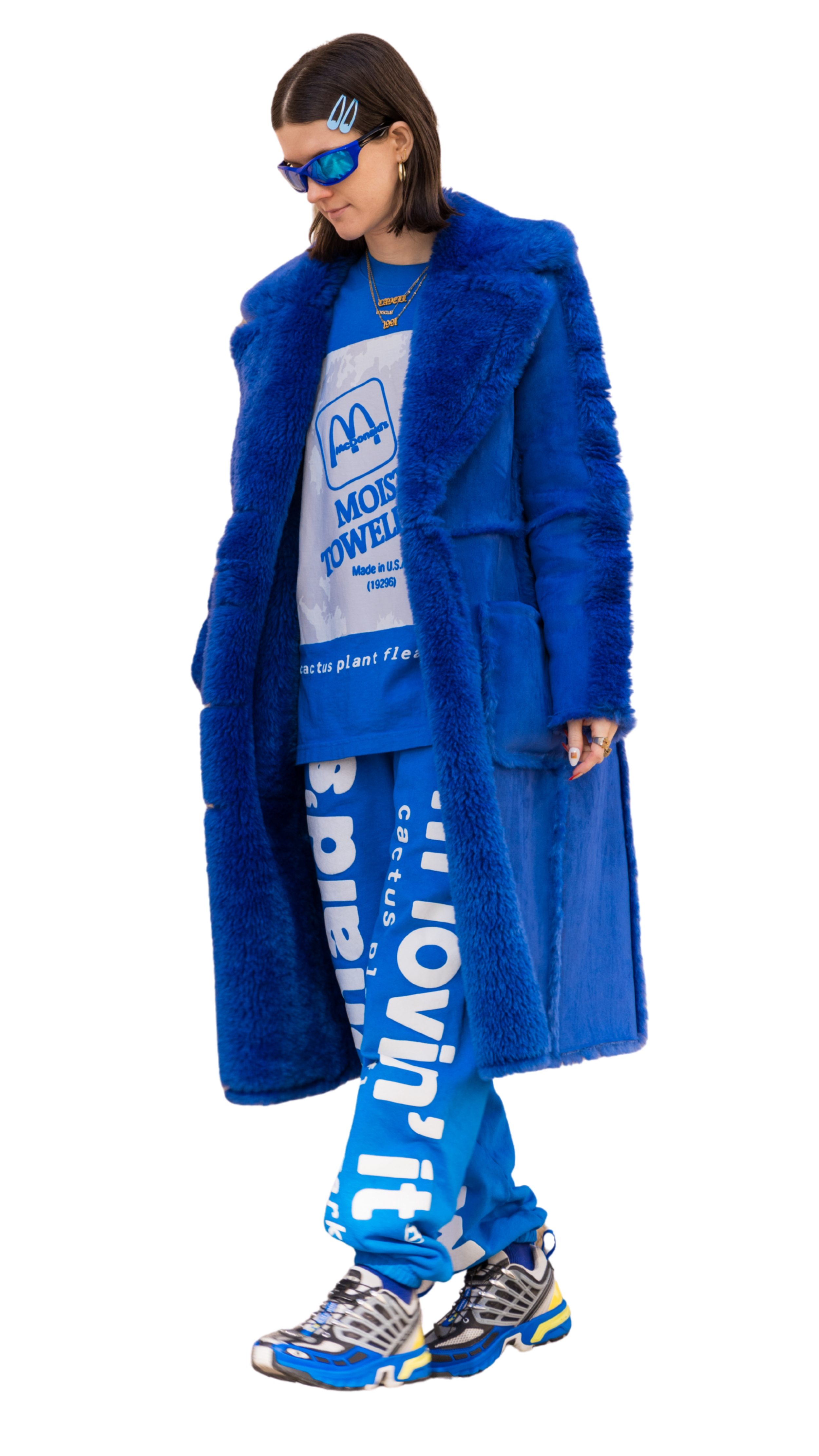 A woman wearing a blue faux fur coat, blue sunglasses, multiple gold necklaces, and printed blue sweatpants and sneakers. She has brown hair with hair clips.