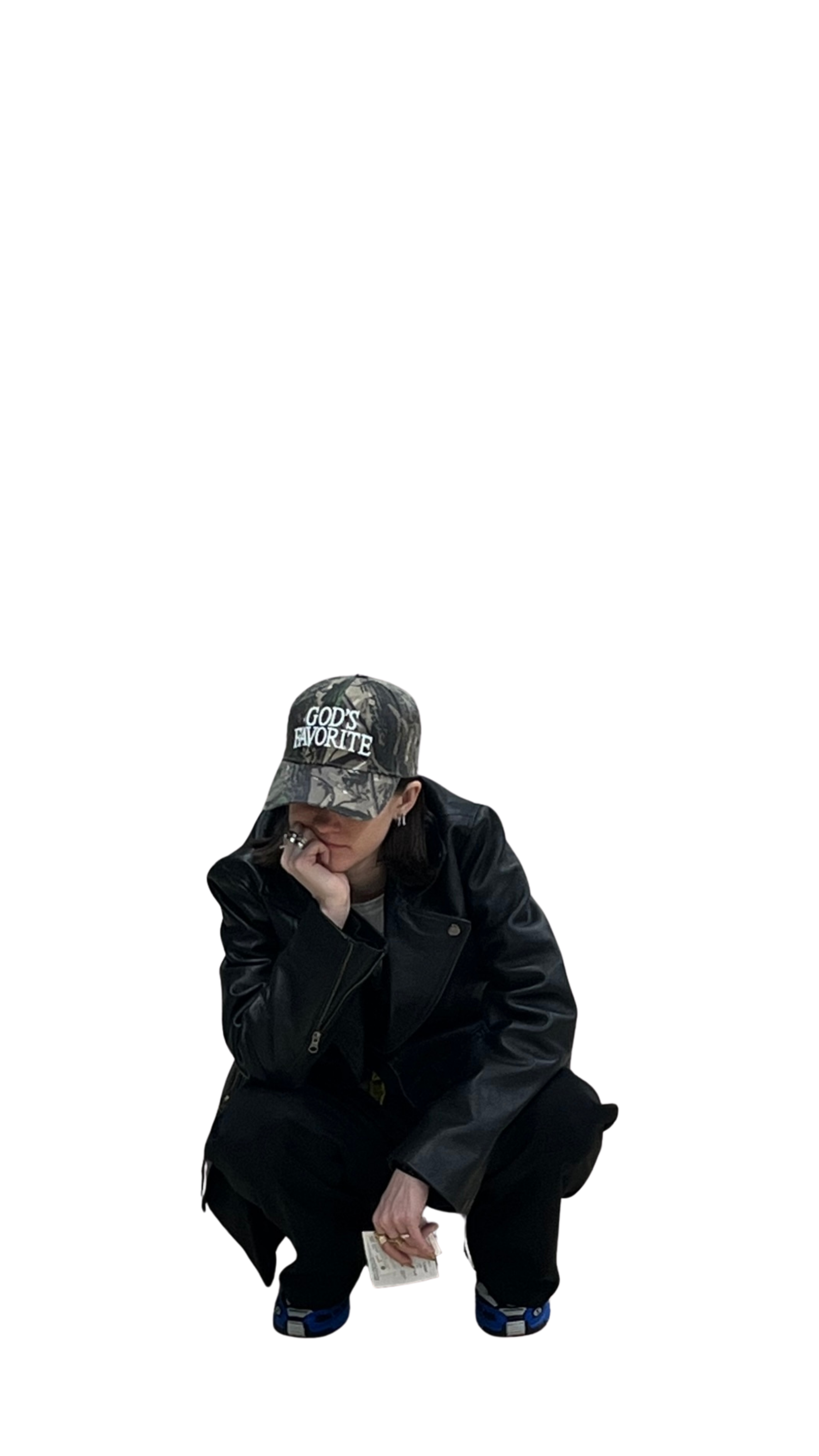 Person wearing a camouflage baseball cap with the words 'GOD'S FAVORITE', a black leather jacket, glasses, and black pants, sitting and resting their chin on their hand while holding a small book or notebook.