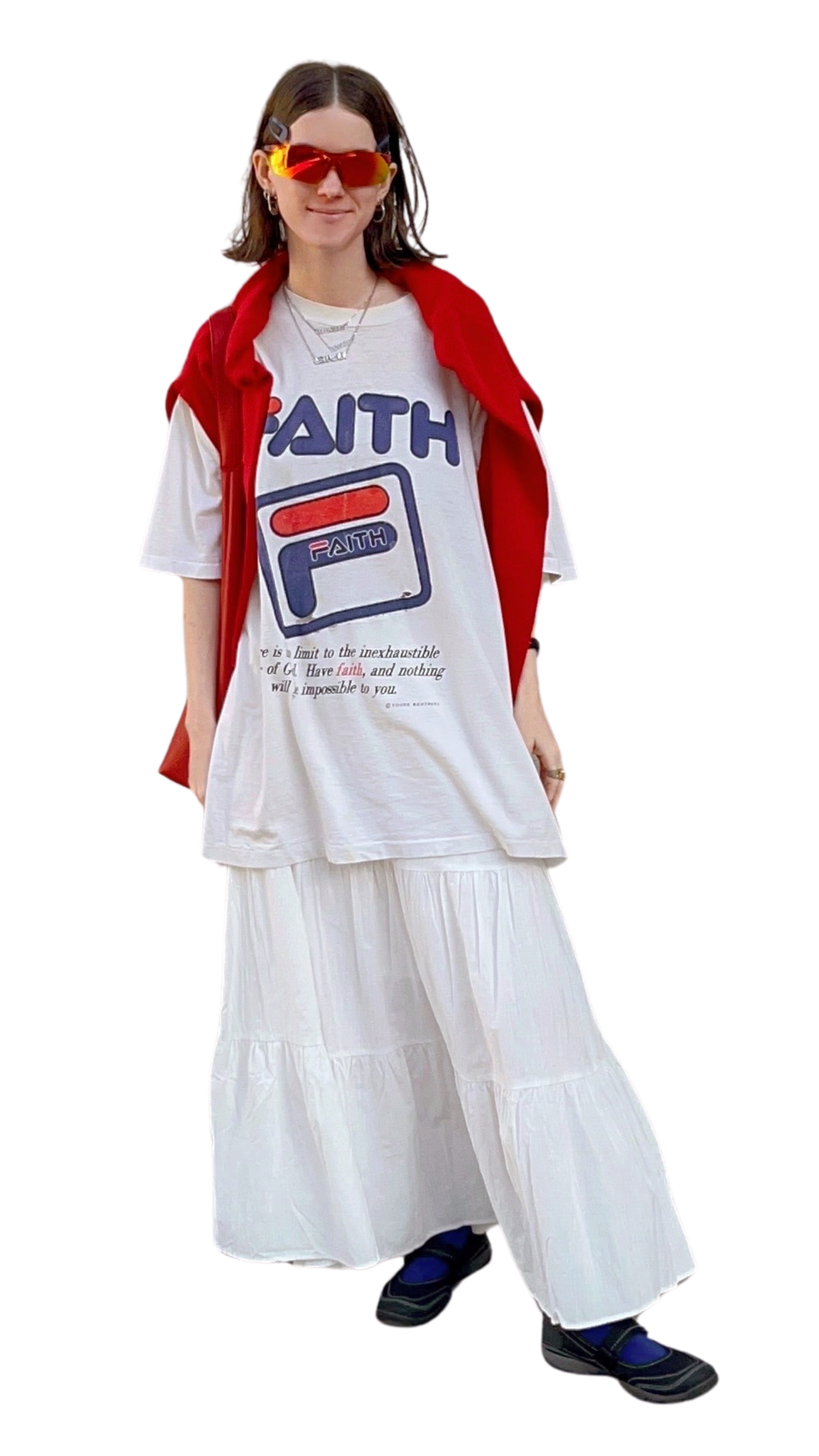 A woman with shoulder-length brown hair wears large red sunglasses, hoop earrings, a white T-shirt with a graphic and text, and a red jacket draped over her shoulders. She also wears a layered necklace and a white pleated skirt with multiple tiers. She is standing against a plain background.