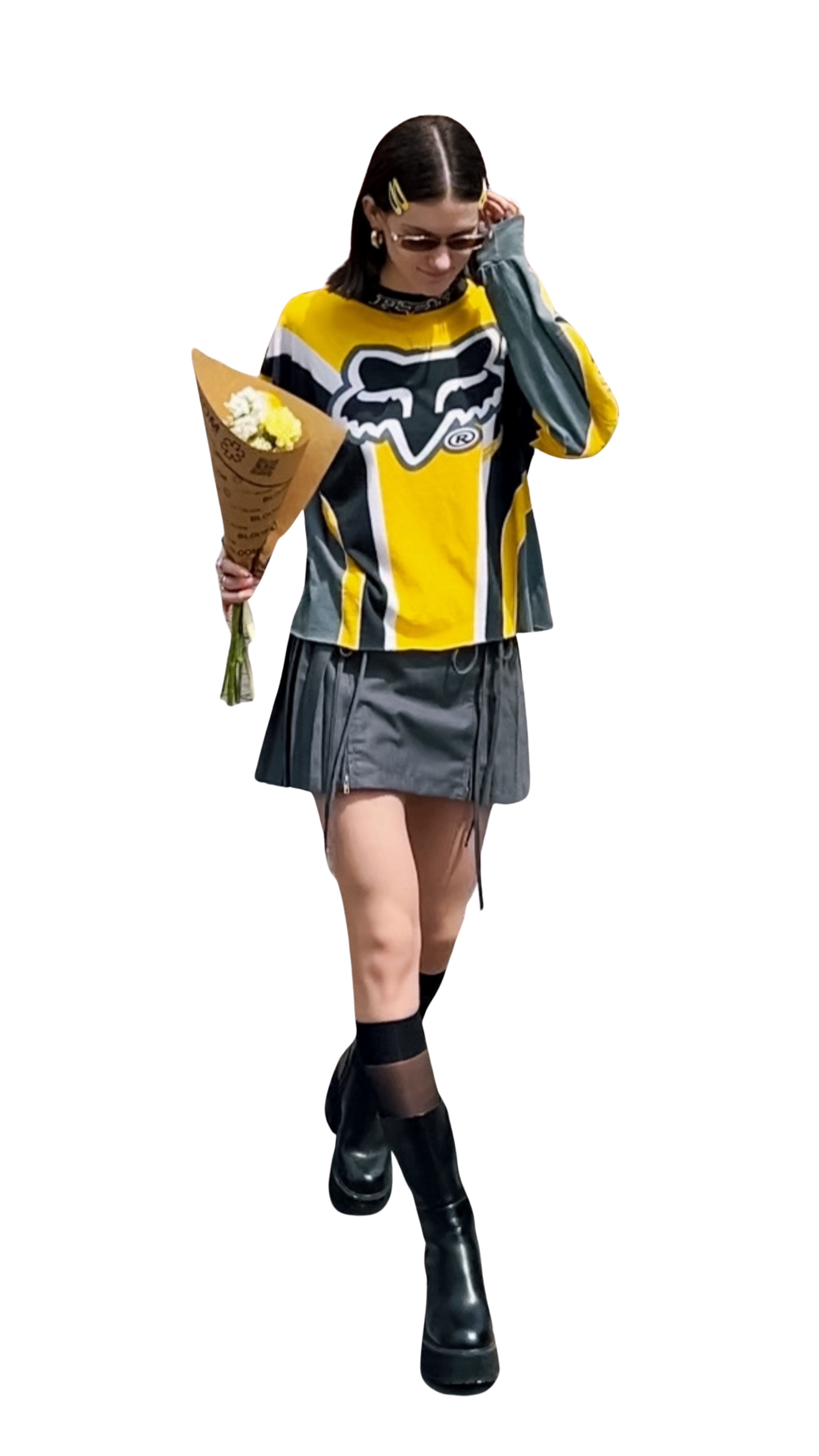 A woman wearing a yellow and black Fox racing motocross jersey, gray mini skirt, black knee-high socks, and black platform boots, holding a bouquet of yellow and white flowers, adjusting her sunglasses while walking.