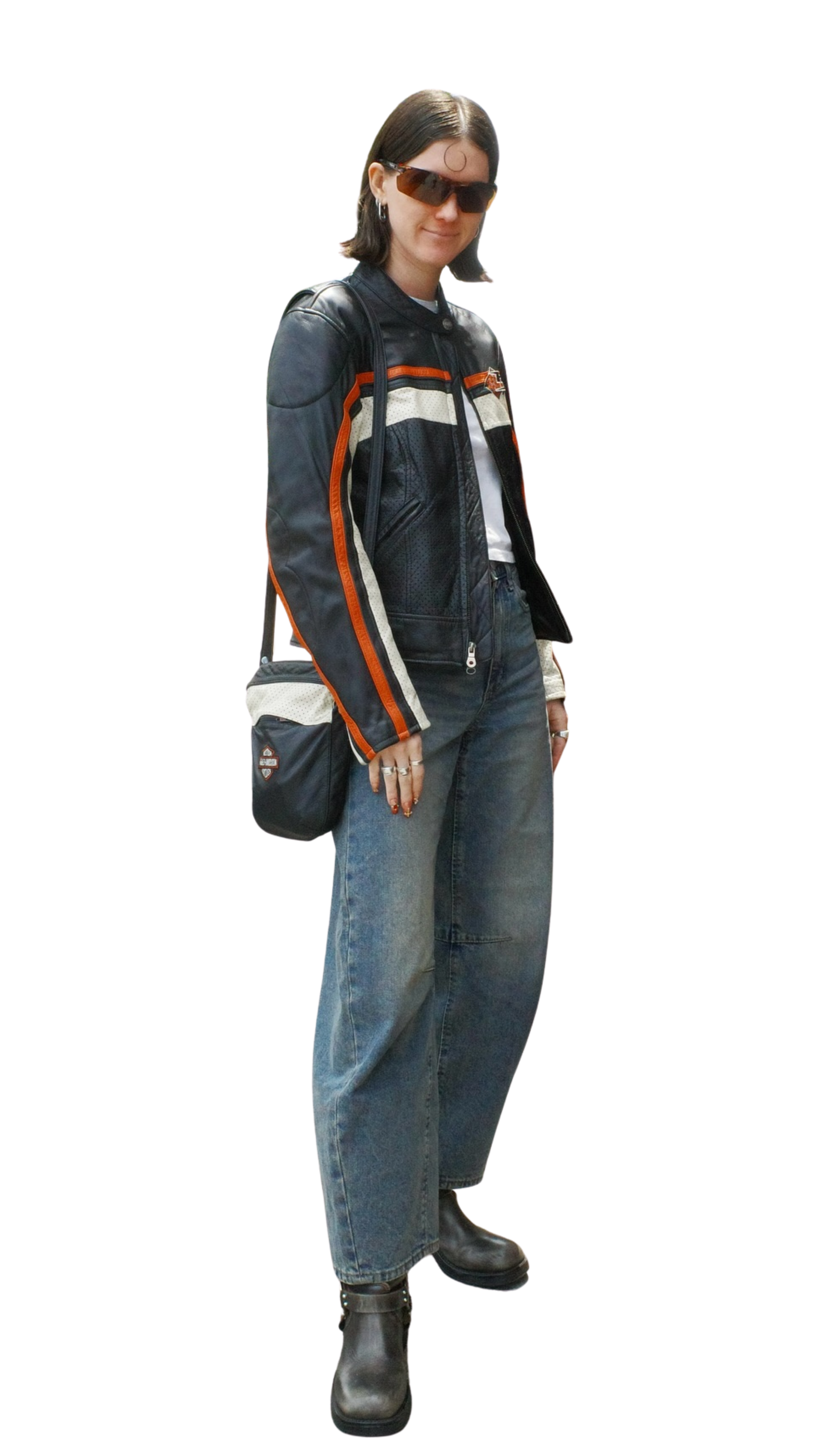 A woman wearing large sunglasses, a leather motorcycle jacket, loose jeans, and black boots, standing with her hands at her sides.