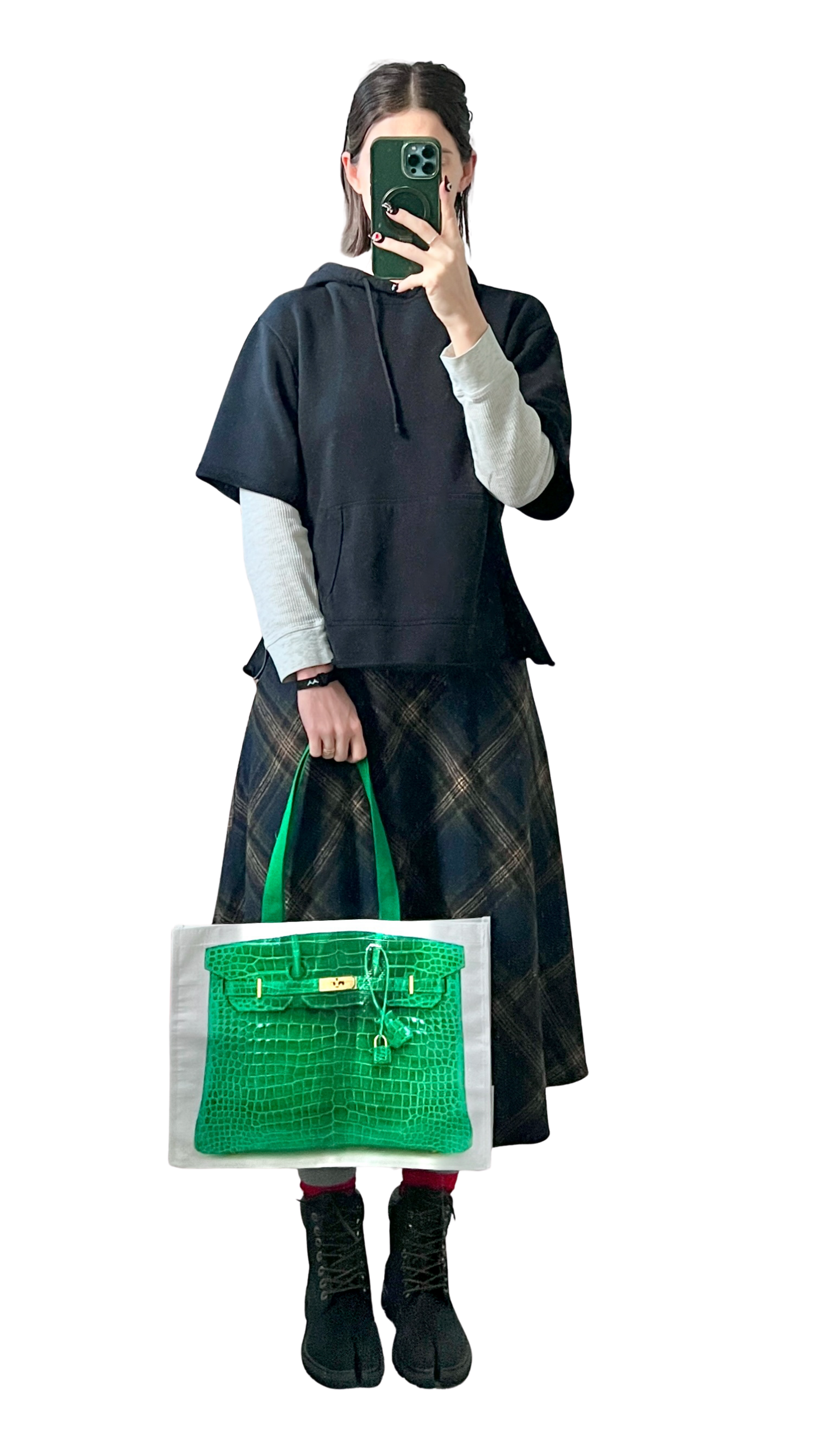 A person taking a selfie while holding a white shopping bag with green accents. They are wearing a black short-sleeved hoodie over a white long-sleeved shirt, a long plaid skirt, black boots, and are standing against a black background.