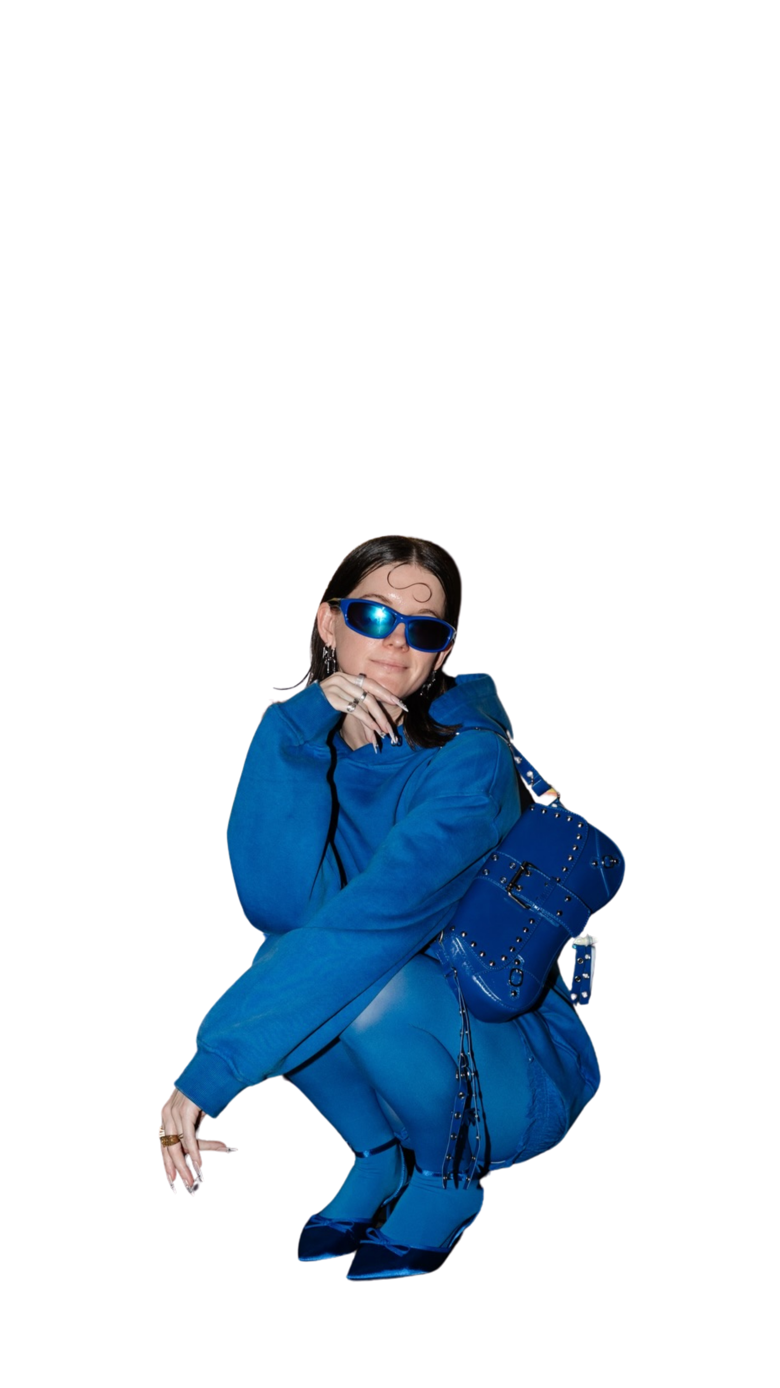 A woman dressed in a blue tracksuit with matching blue accessories, including sunglasses, a handbag, and shoes, is crouching down against a black background.