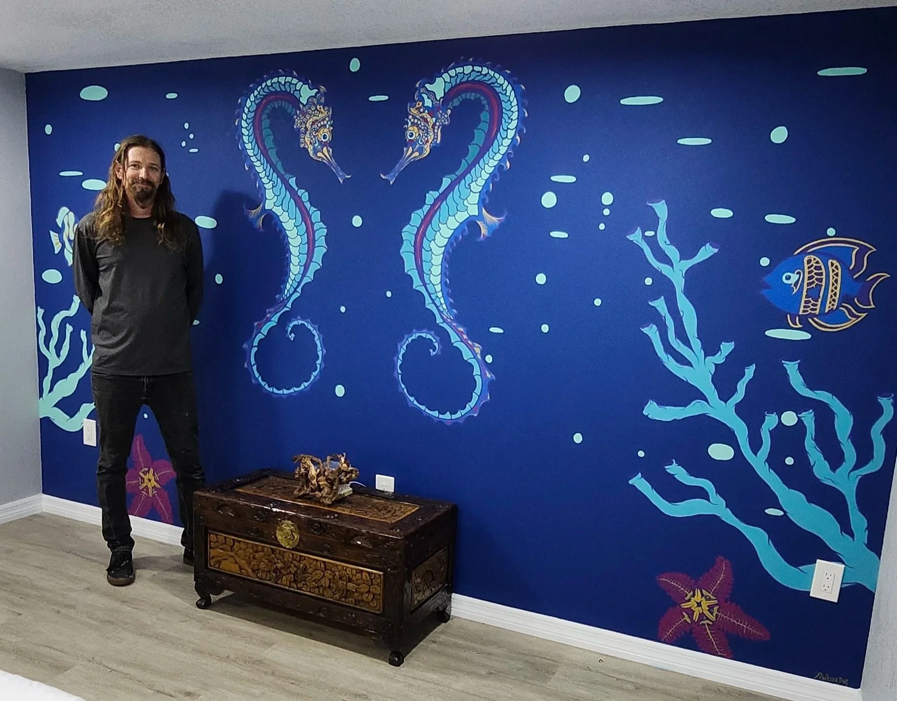 Madhava Das standing in front of a vibrant blue underwater-themed mural featuring two seahorses, a fish, seaweed, and coral.