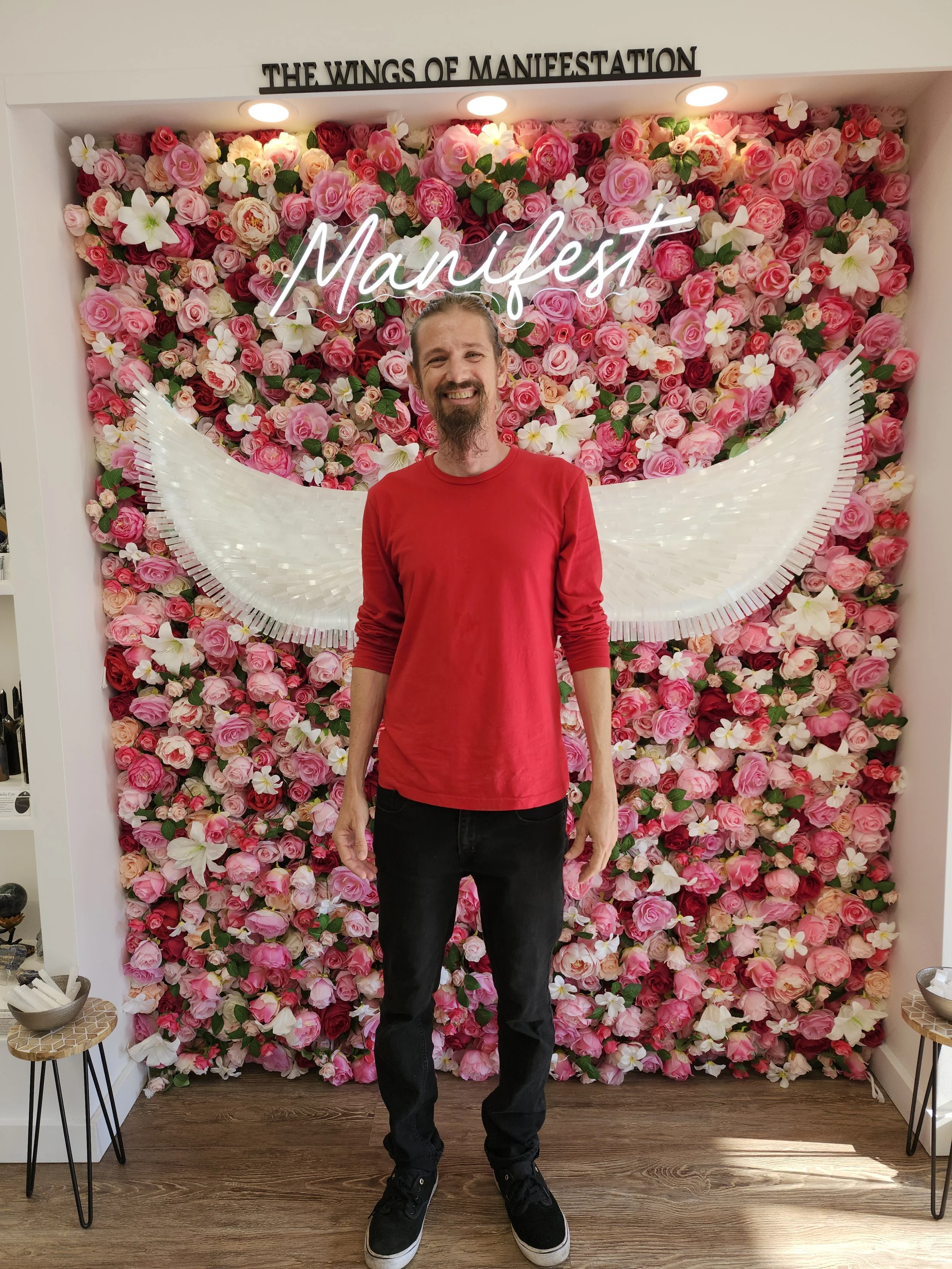 A man with long hair and a beard wearing a red sweater and black pants standing in front of a floral wall with pink and white flowers. There are white angel wings decorative art behind him and a neon sign that reads 'Manifest'.