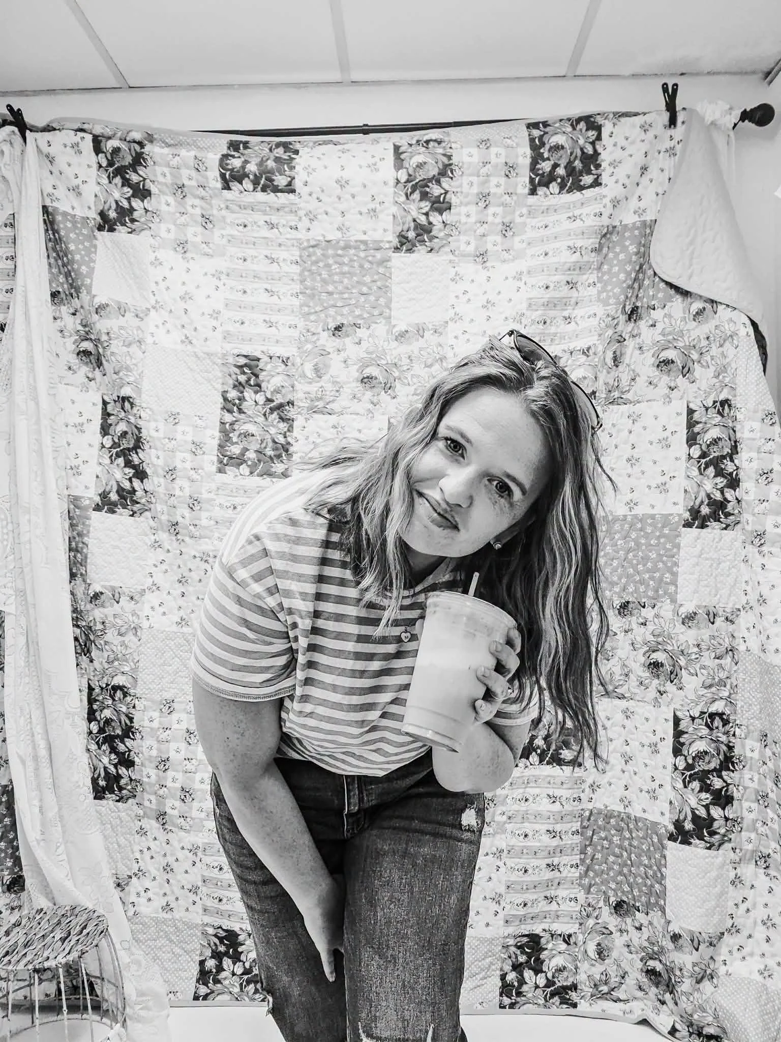 A woman with wavy hair and sunglasses on her head is holding a drink and leaning against a quilted floral fabric backdrop, smiling at the camera.