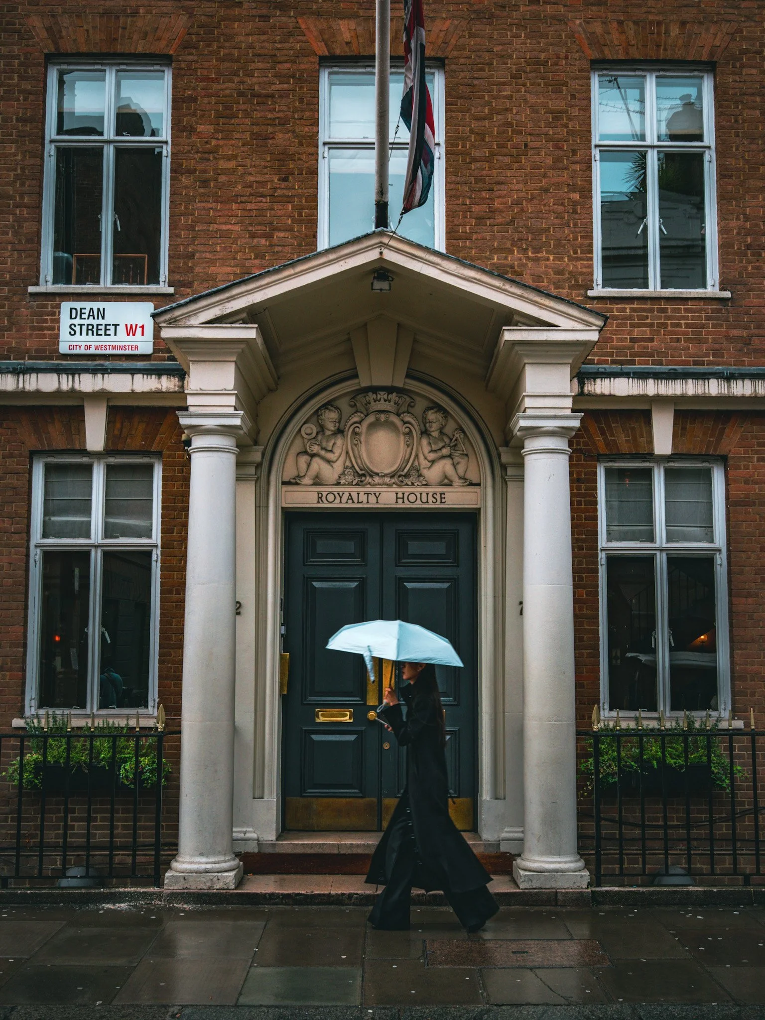 A woman in a long black coat walking in front of a building with white columns and a sign that reads 'Royalty House' on a rainy day, holding a light blue umbrella.