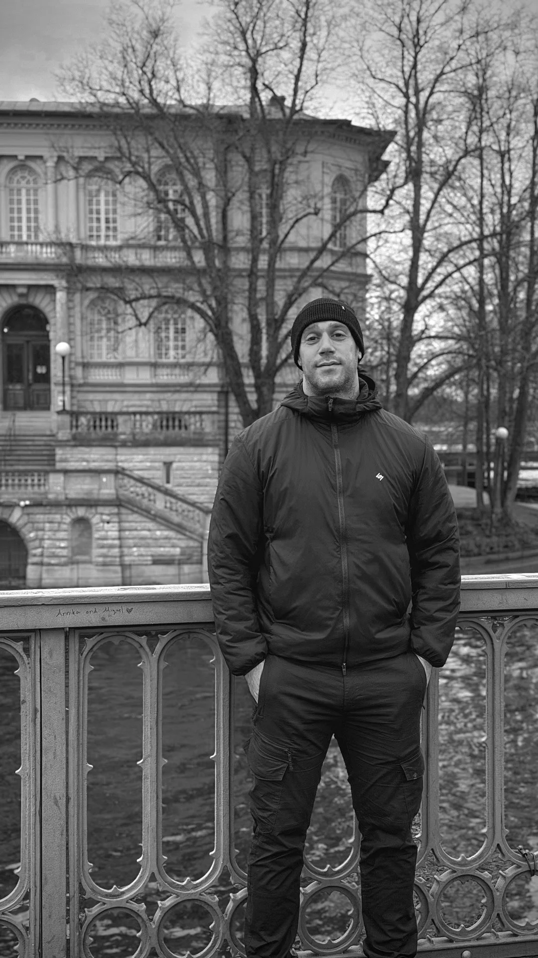 A man stands on a bridge in front of a historic building with large windows and trees without leaves, in a black-and-white photo.