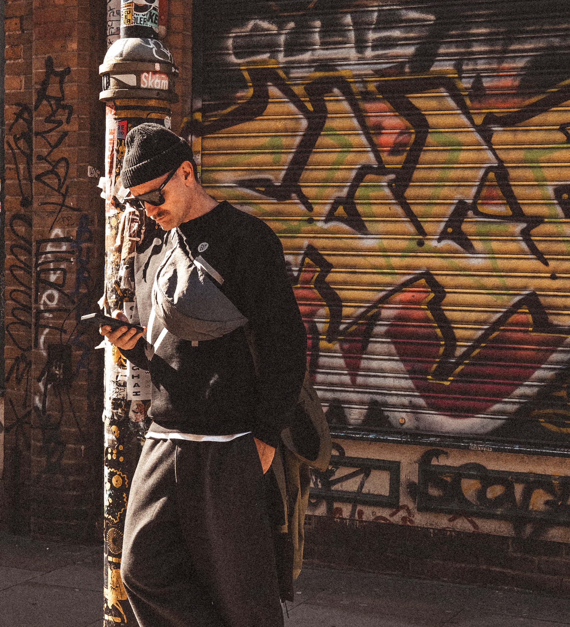 A man in a black beanie, glasses, and a gray jacket looking at his phone while standing in front of a graffiti-covered rolling metal door and brick wall.