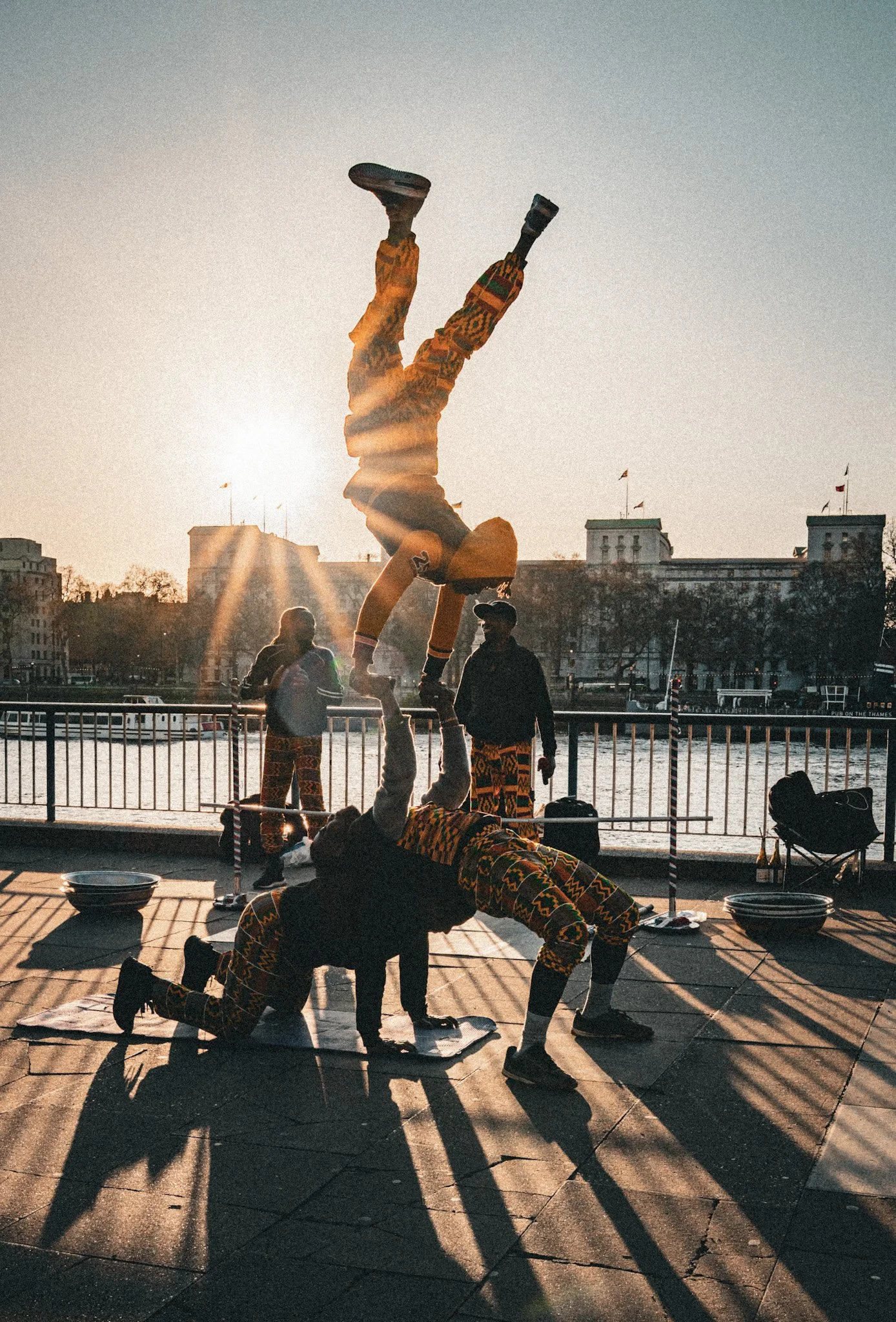 People performing acrobatic stunts and yoga poses on a dock near water during sunset, with city buildings and flags in the background.