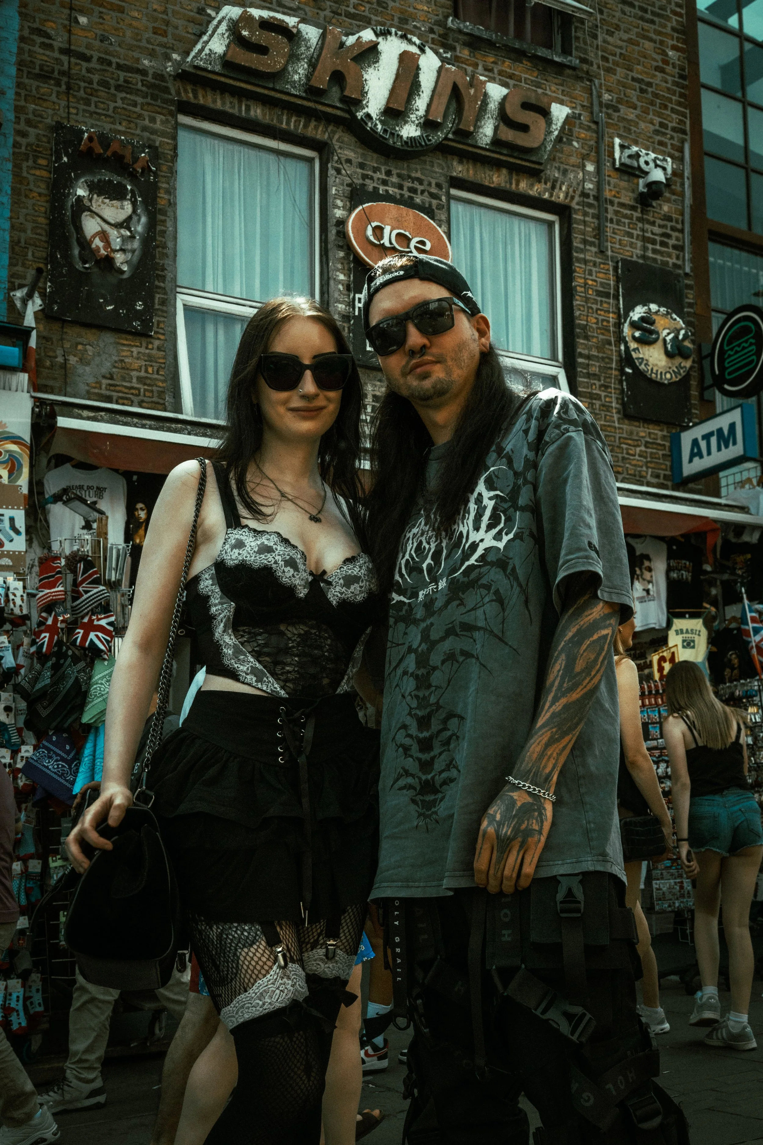 Two young adults, a woman and a man, standing in front of a brick building with signs for a 'Skins' store and an 'Ace' store. The woman is wearing sunglasses, a black lace top, fishnet stockings, and carrying a black purse. The man is wearing sunglas