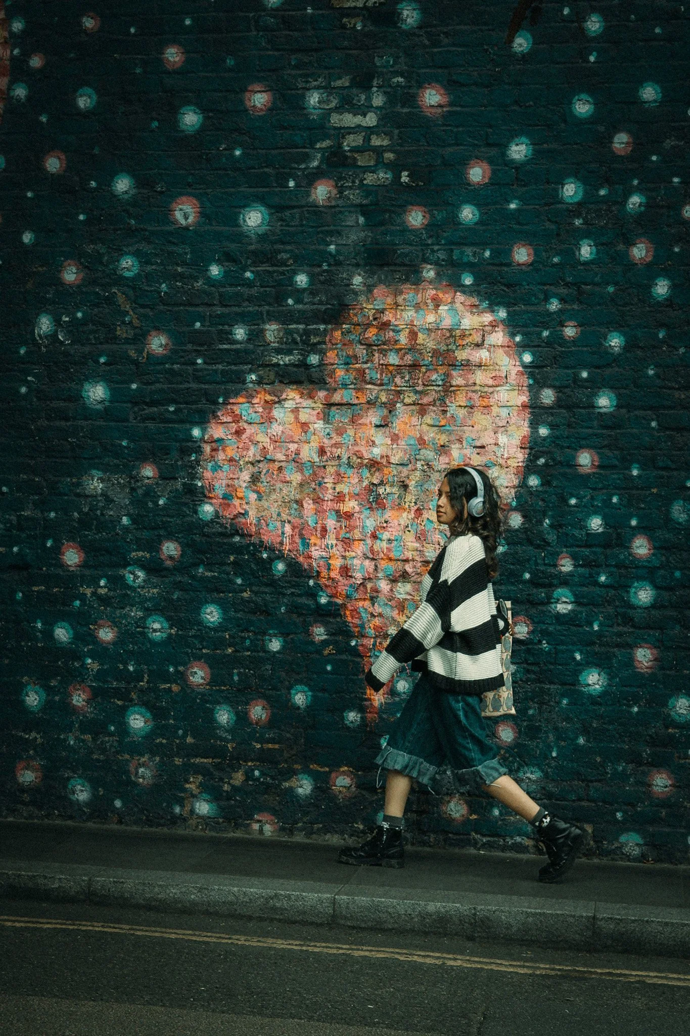 A woman walking past a large graffiti mural of a girl's face on a brick wall, wearing headphones, a black and white striped sweater, a long skirt, and combat boots.