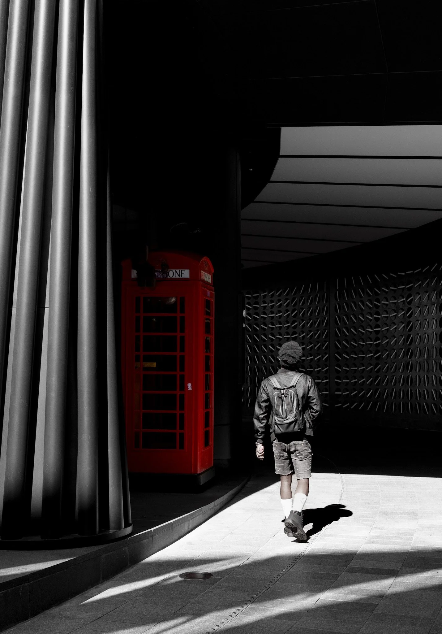 A person walking towards an architectural structure with a geometric pattern and lighting that creates a high-contrast scene. The person is wearing a backpack, shorts, and sneakers, and is casting a shadow on the ground. There's a traditional red Bri