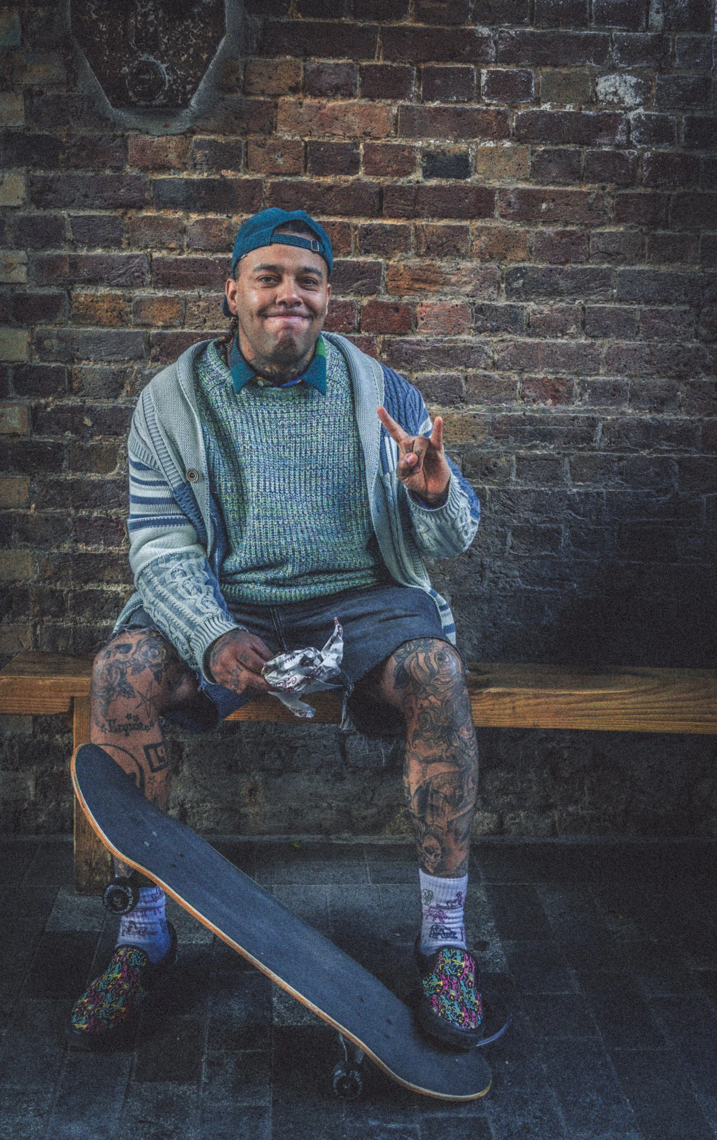 A young man with tattoos on his arms and legs, wearing a cap backwards, colorful socks, and sneakers, sitting on a wooden bench in front of a brick wall. He is holding a skateboard and making a peace sign with his hand, smiling at the camera.