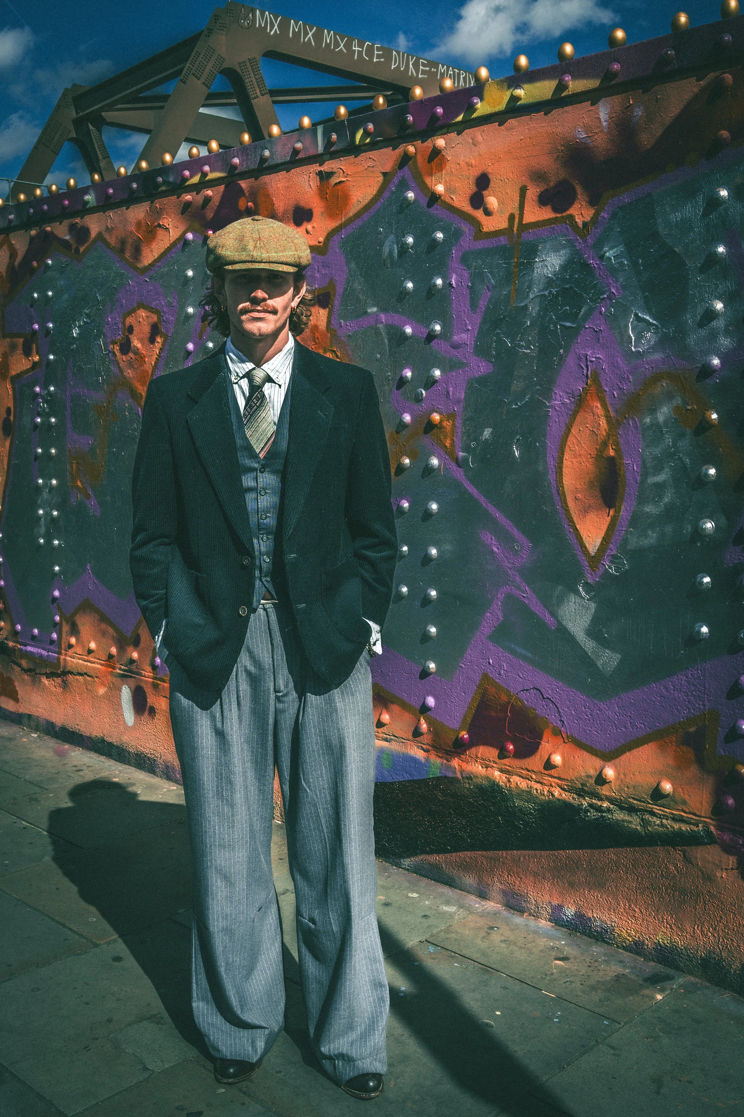 Man with a mustache and curly hair wearing a vintage-style flat cap, a black pinstripe suit, a patterned vest, a short-sleeve shirt, a tie, and wide-leg trousers, standing against a colorful, graffiti-covered wall with metallic studs, under a cloudy 