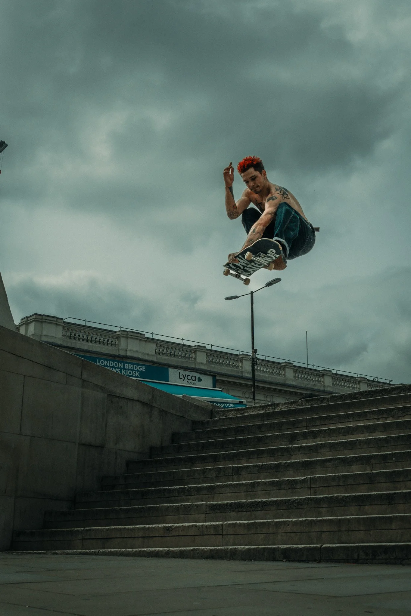 A shirtless man with red hair and tattoos performing a skateboard trick in a city setting, with steps in the foreground and a cloudy sky above.