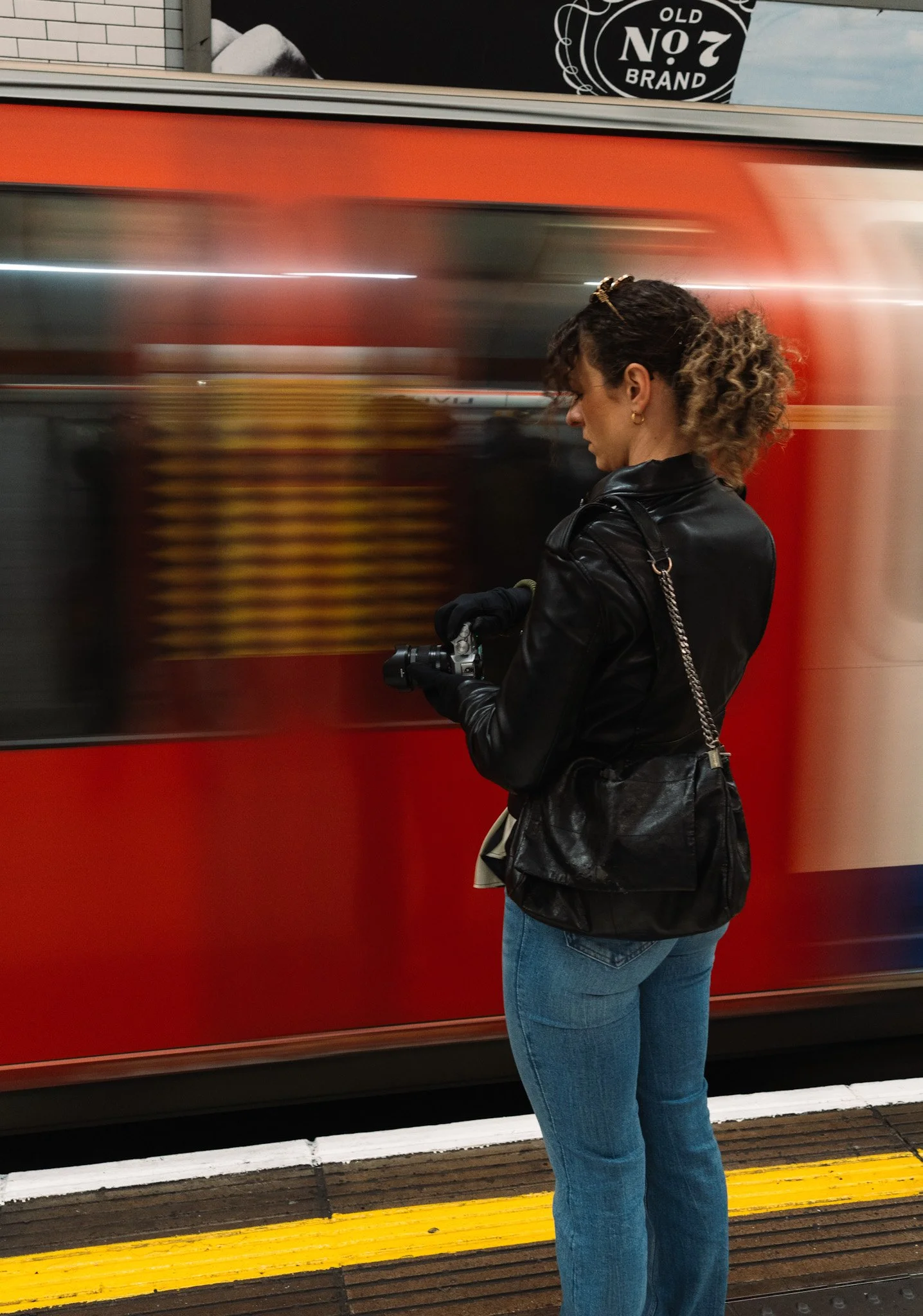 A woman with curly hair, wearing a black leather jacket and blue jeans, stands on a train platform looking at her phone as a red train passes by.