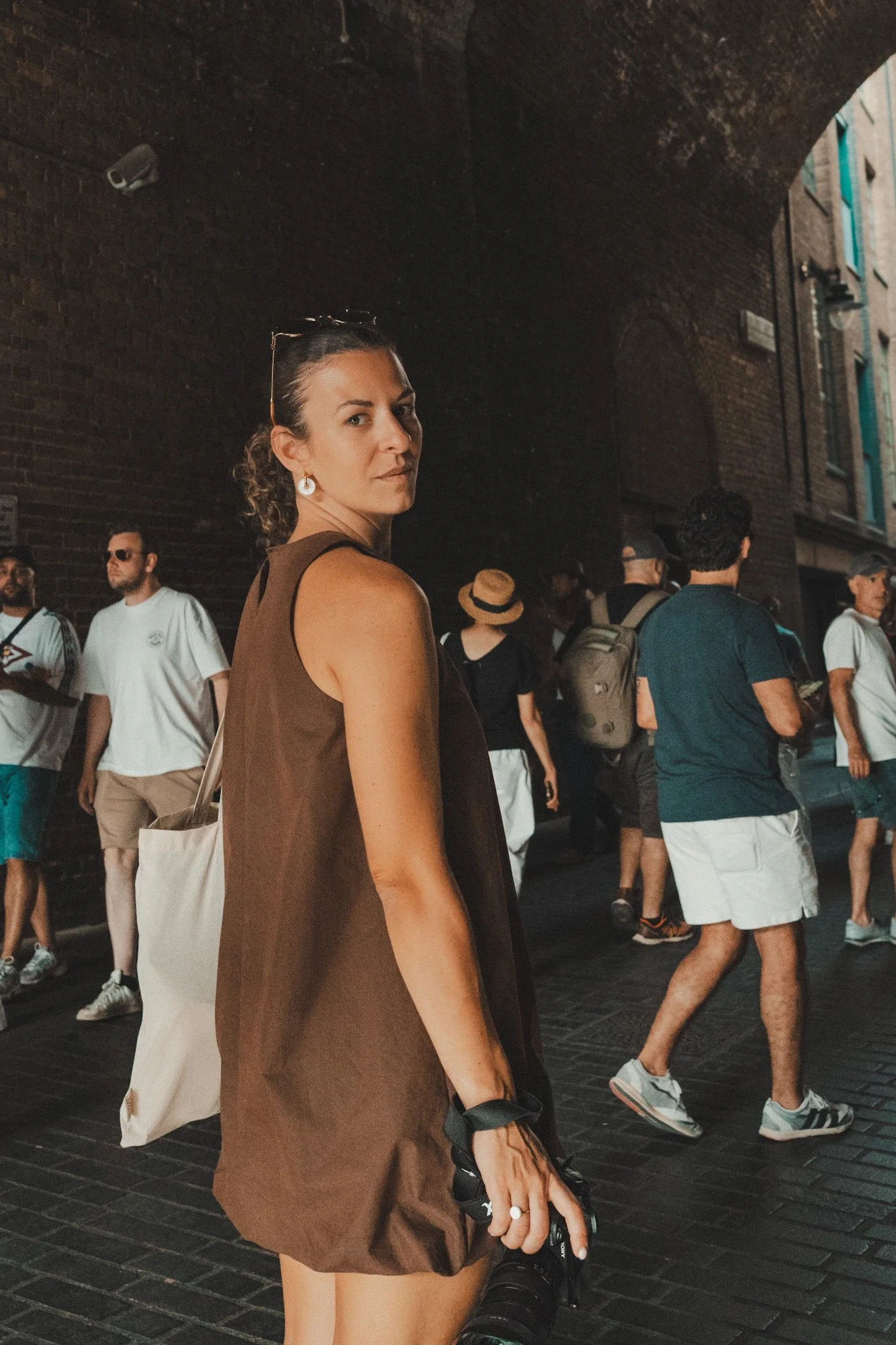 A woman in a brown sleeveless dress holding a camera, standing in a crowd on a city street, with a dark brick wall and other pedestrians in the background.