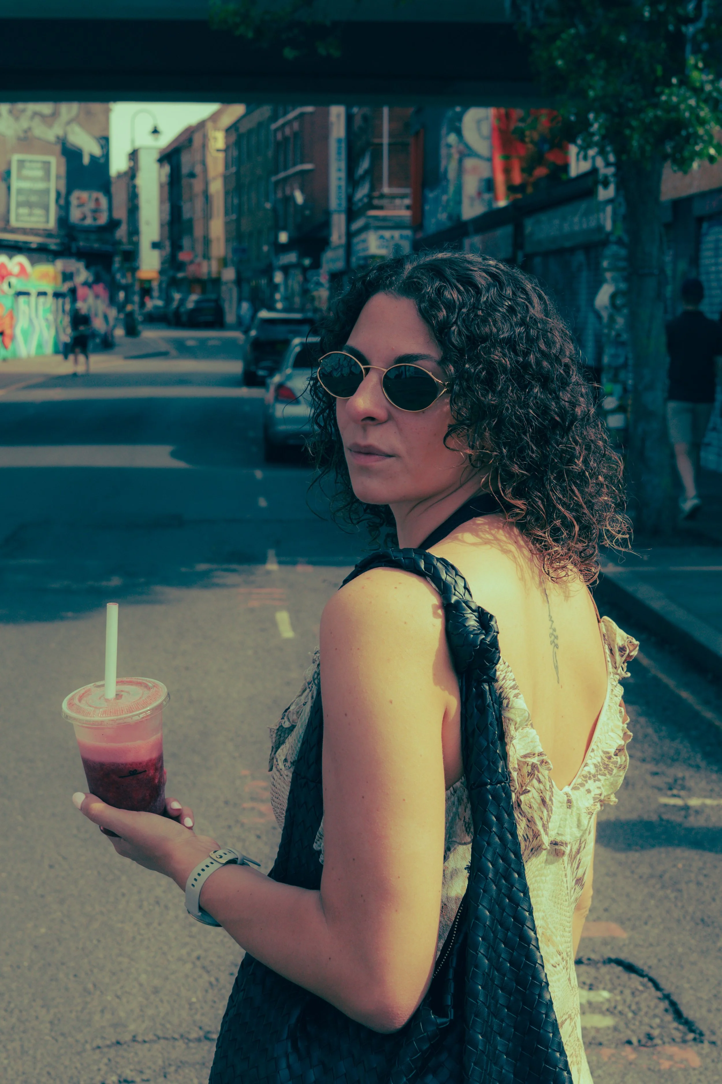 A woman with curly hair wearing sunglasses and a yellow dress, holding a pink layered drink with a straw, standing on a city street.