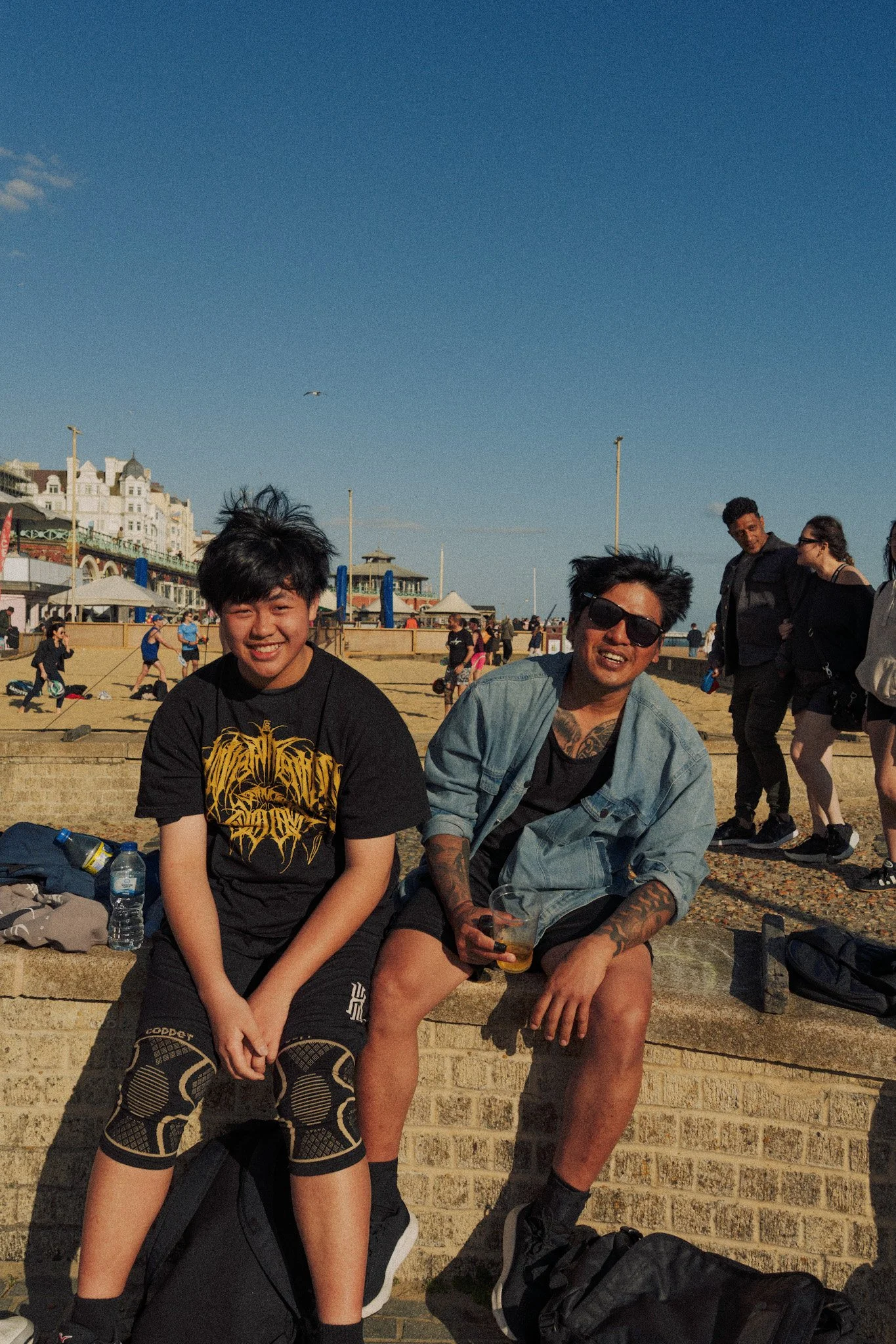 Two young men sitting on a brick wall at the beach, smiling and enjoying the sunny day. One is wearing a black t-shirt with a lion design, the other is in a denim jacket and sunglasses. In the background, people are walking, relaxing, and playing at 