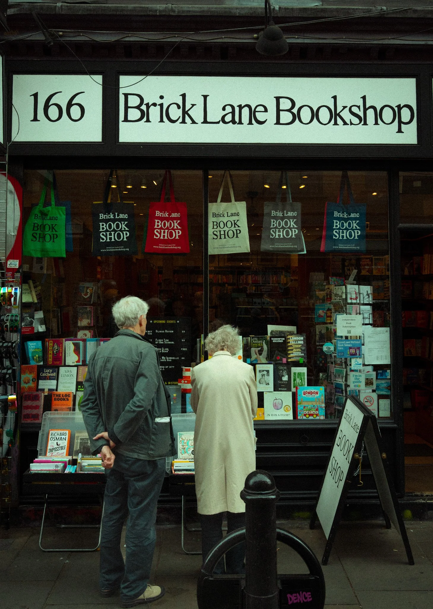 A storefront for Brick Lane Bookshop at 166 with several people browsing outside. The shop has a large sign with the shop's name and number, and colorful tote bags with the shop's name hanging in the window. Inside, shelves filled with books are visi