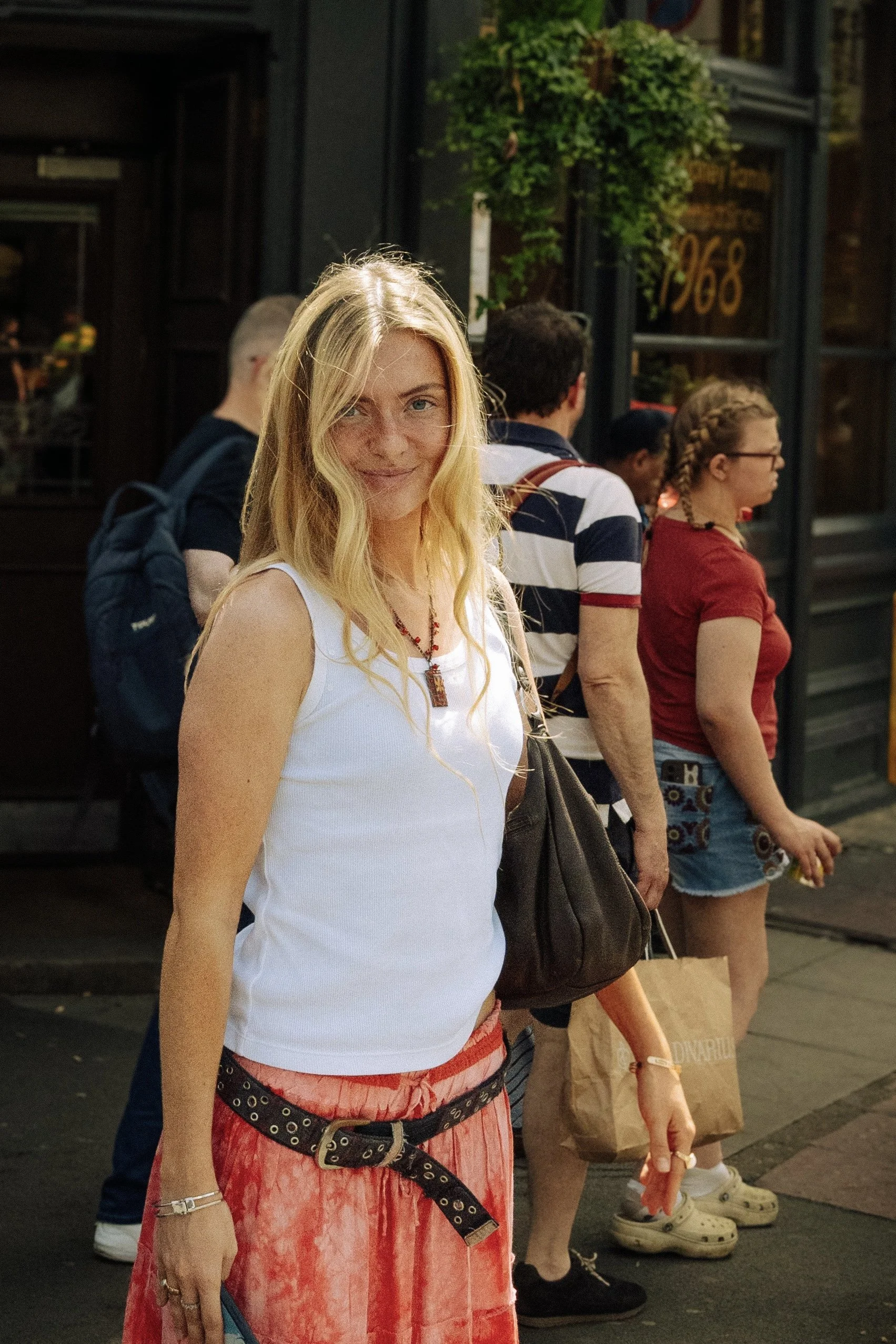 A blonde woman in a white tank top and long pink patterned skirt stands in front of a crowd of people outside a building with green plants in the window. The people are waiting in line or walking past, and she is smiling at the camera.