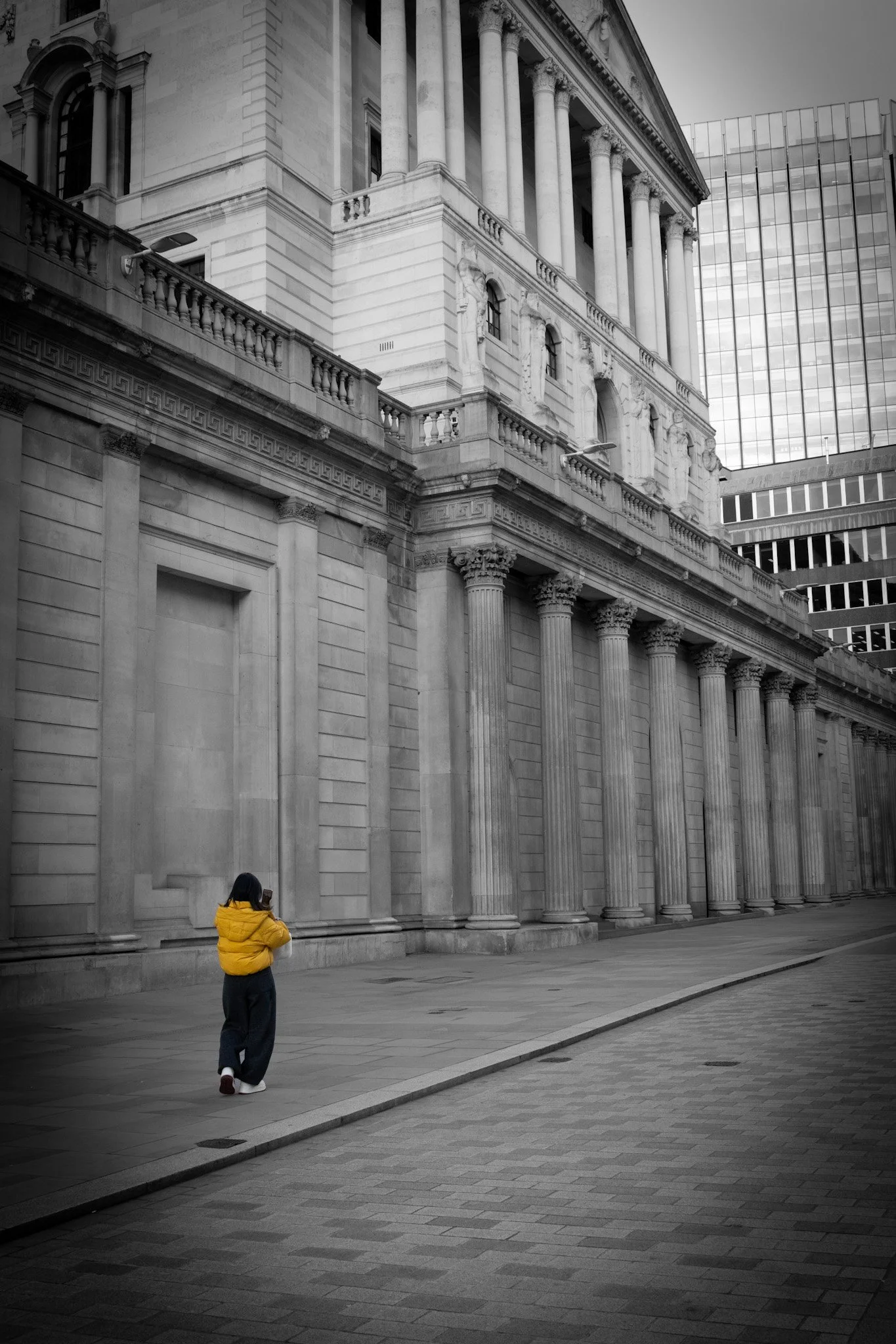 A person wearing a bright yellow jacket standing on a city sidewalk in front of a large, historic building with columns and ornate architectural details, partly in black and white with a modern glass building in the background.