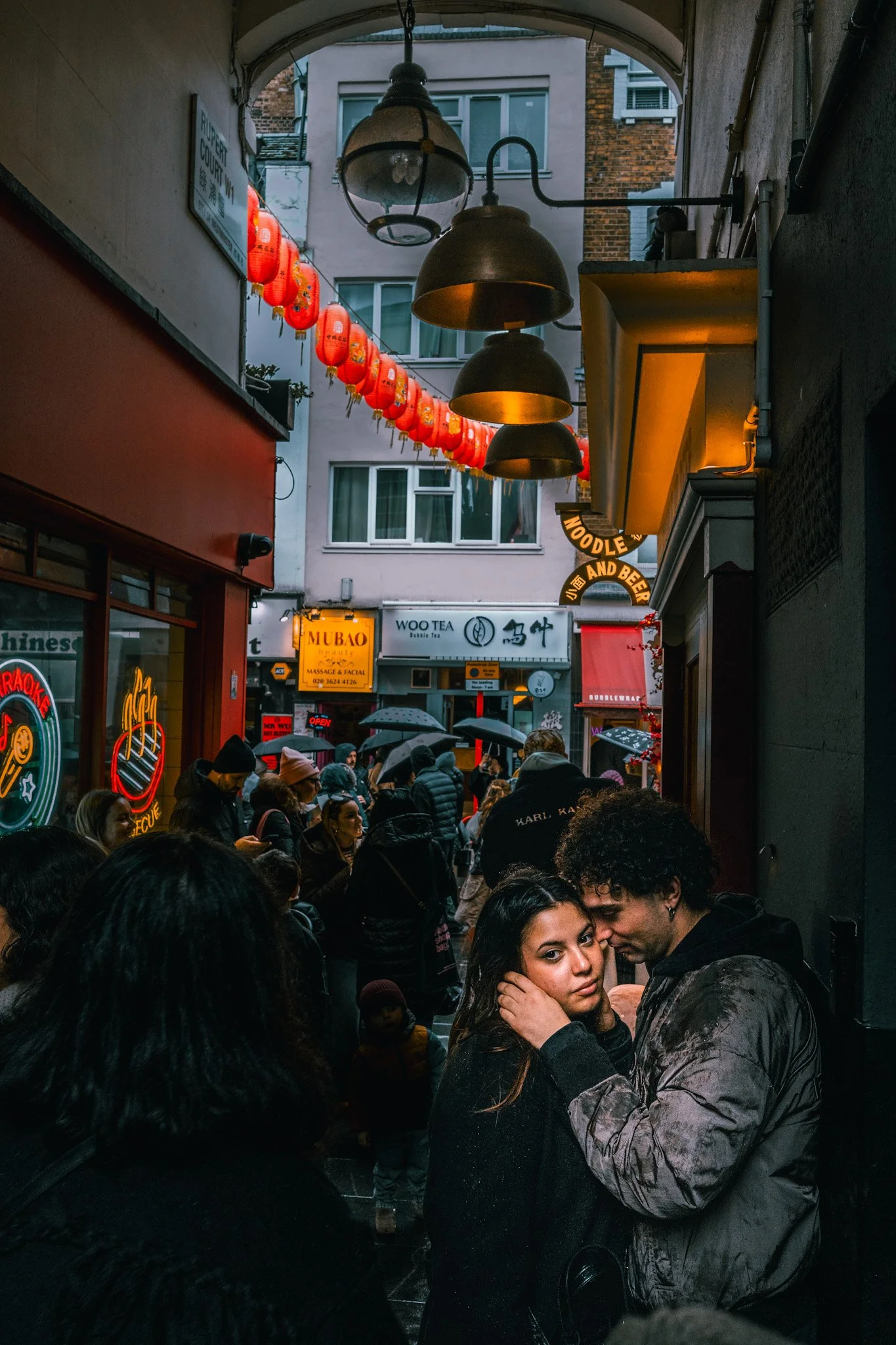 Crowded alleyway with people under red Chinese lanterns, some holding umbrellas, and two people embracing in the foreground.