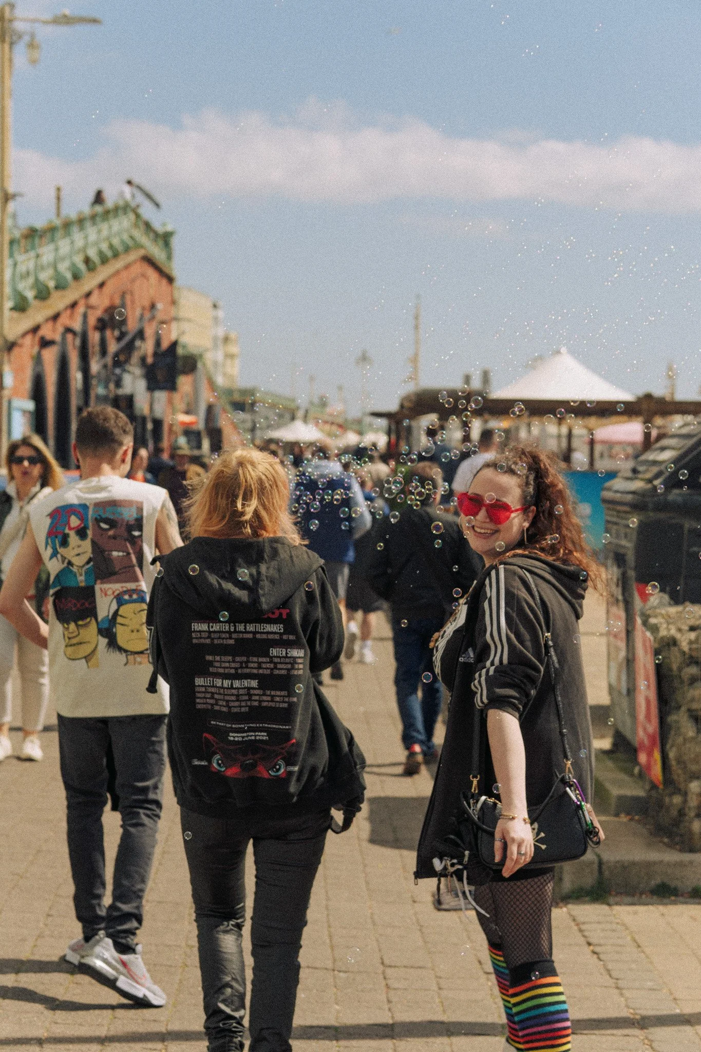 People walking outdoors at a busy fair or market on a sunny day, with a woman smiling and wearing rainbow striped thigh-high socks and red sunglasses, surrounded by others in casual clothing.