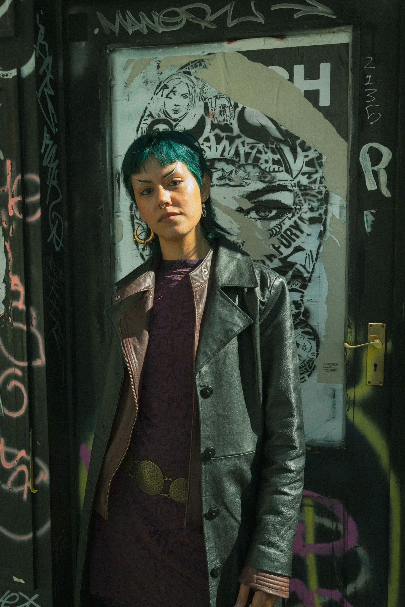 A woman with short black hair with teal streaks and a septum piercing, wearing a black leather jacket and a purple lace dress with a gold chain belt, standing in front of a graffiti-covered door with a large black and white portrait of a woman's face